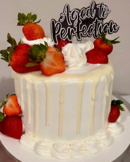 A white birthday cake with strawberries and whipped cream decorations, topped with a sign that reads "Aged to Perfection."