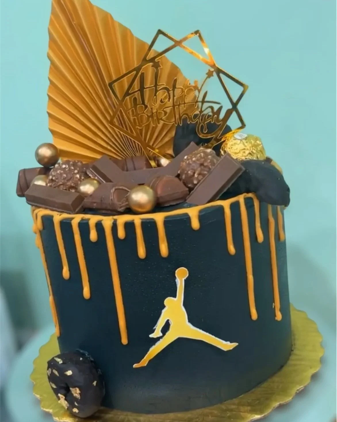 A birthday cake with a black and gold color theme, featuring a Jumpman logo on the front, gold drip decorations, chocolate pieces, gold spheres, and a large gold fan topper with a 'Happy Birthday' sign.