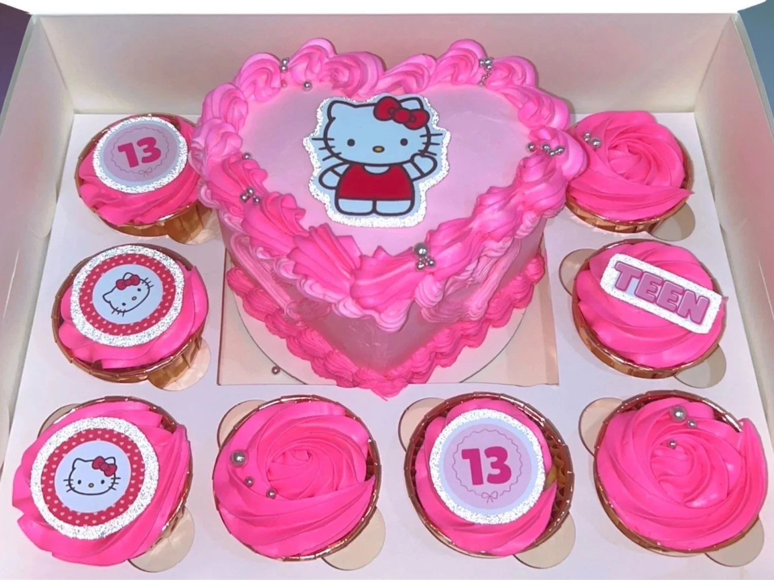 A heart-shaped birthday cake with pink frosting and Hello Kitty decoration in a box, surrounded by eight matching cupcakes with pink frosting and Hello Kitty or '13' decorations.