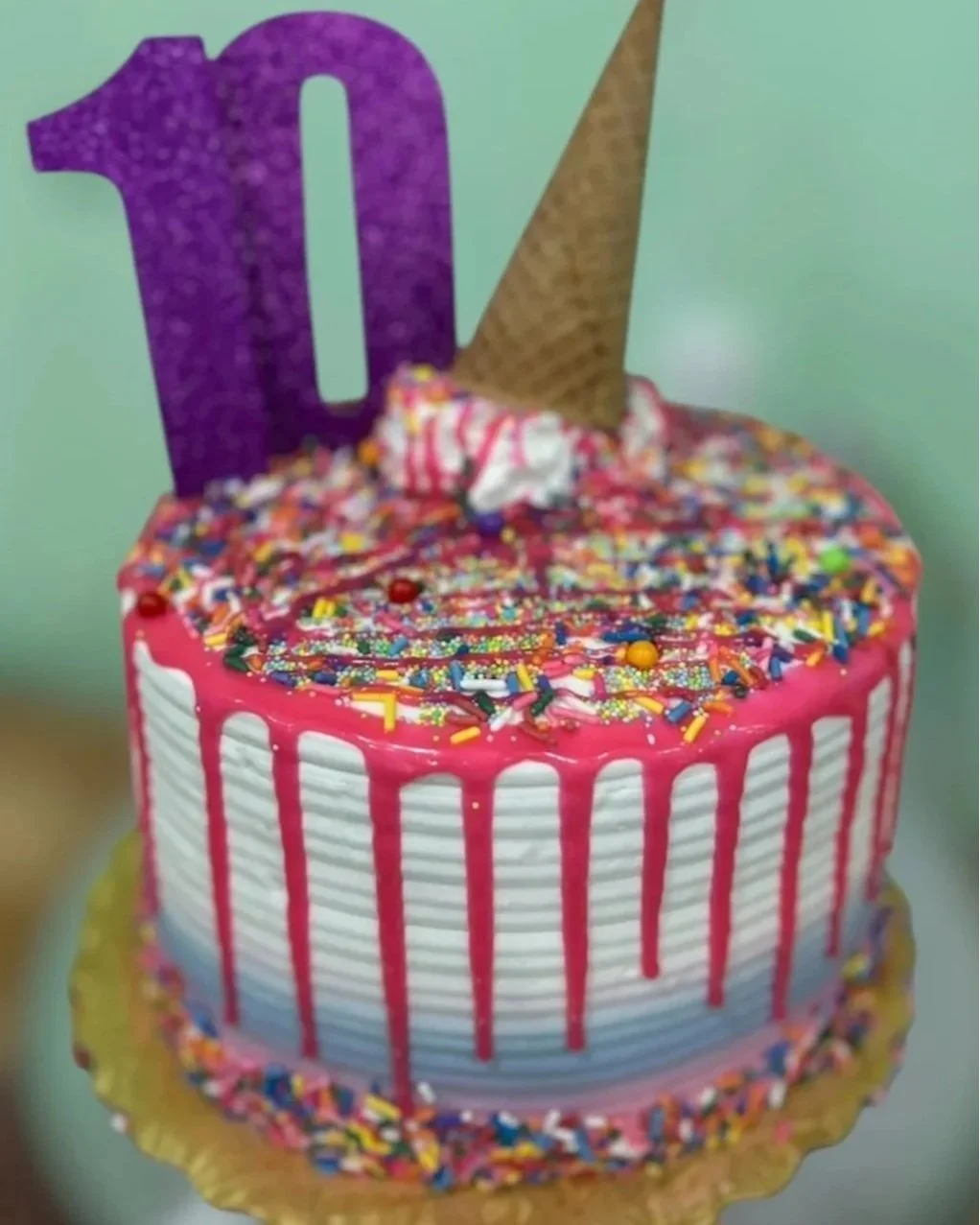 A decorated birthday cake with colorful sprinkles, pink drip icing, and a large purple foam number ten. It also has a waffle cone and a small party hat on top.