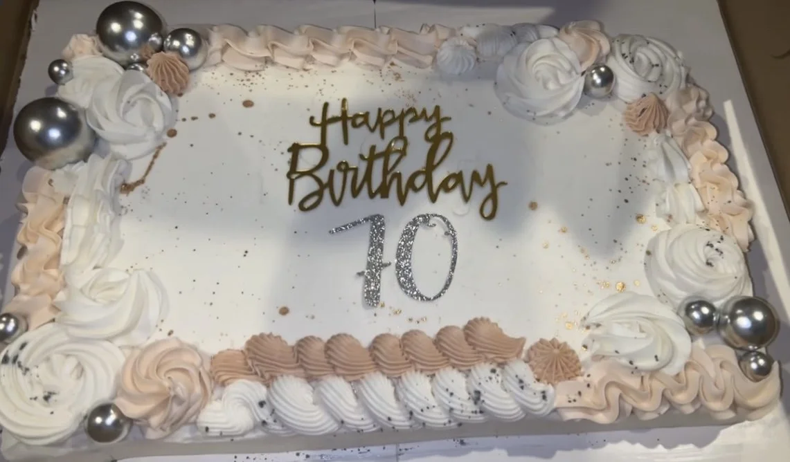 A rectangular birthday cake with white icing, decorated with light pink and white piped swirls and silver edible balls. The cake has a message written in gold and silver icing that reads "Happy Birthday 10."