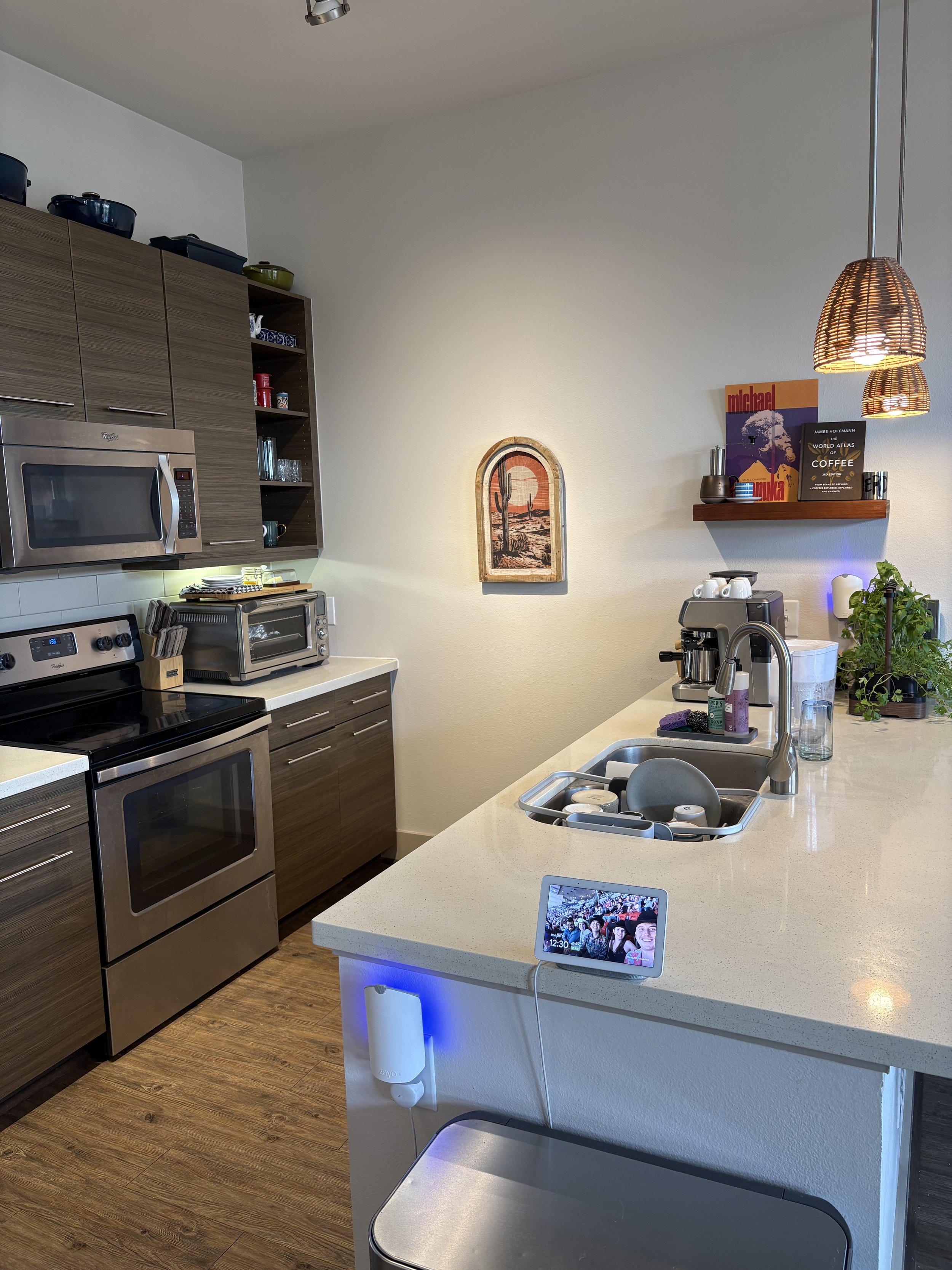 Modern kitchen with wooden cabinets, white countertops, stainless steel appliances, a small tablet displaying a photo, and various kitchen items including a plant, books, and small appliances.