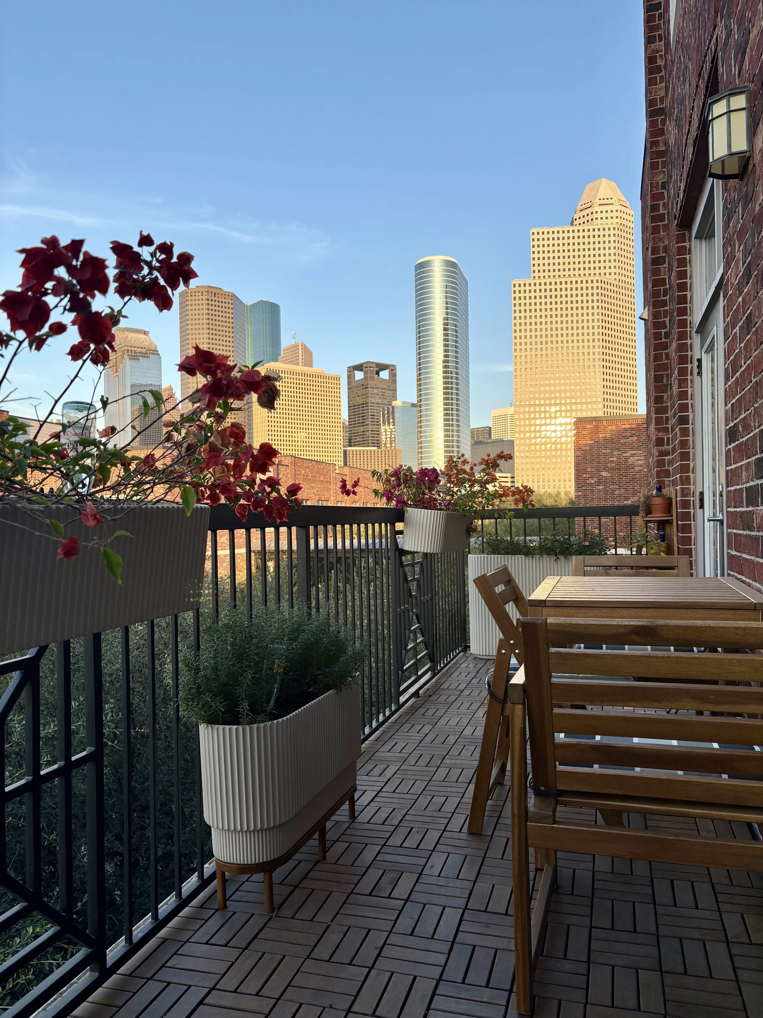 View of a balcony with wooden tables and chairs, potted plants with red and purple flowers, and a city skyline with skyscrapers in the background during sunset.
