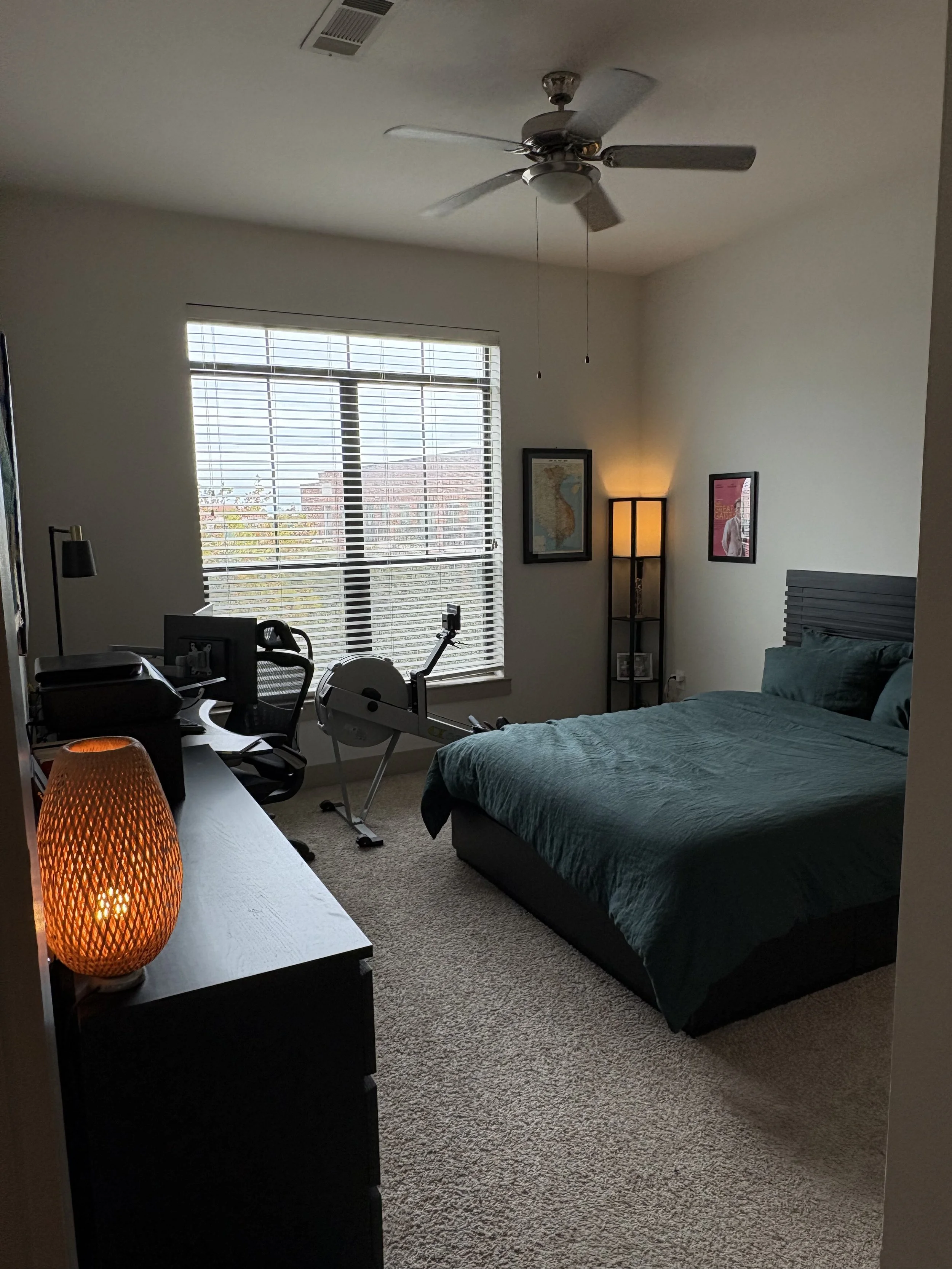 A bedroom with a bed, window, exercise bike, desk with computer, and ambient lighting.