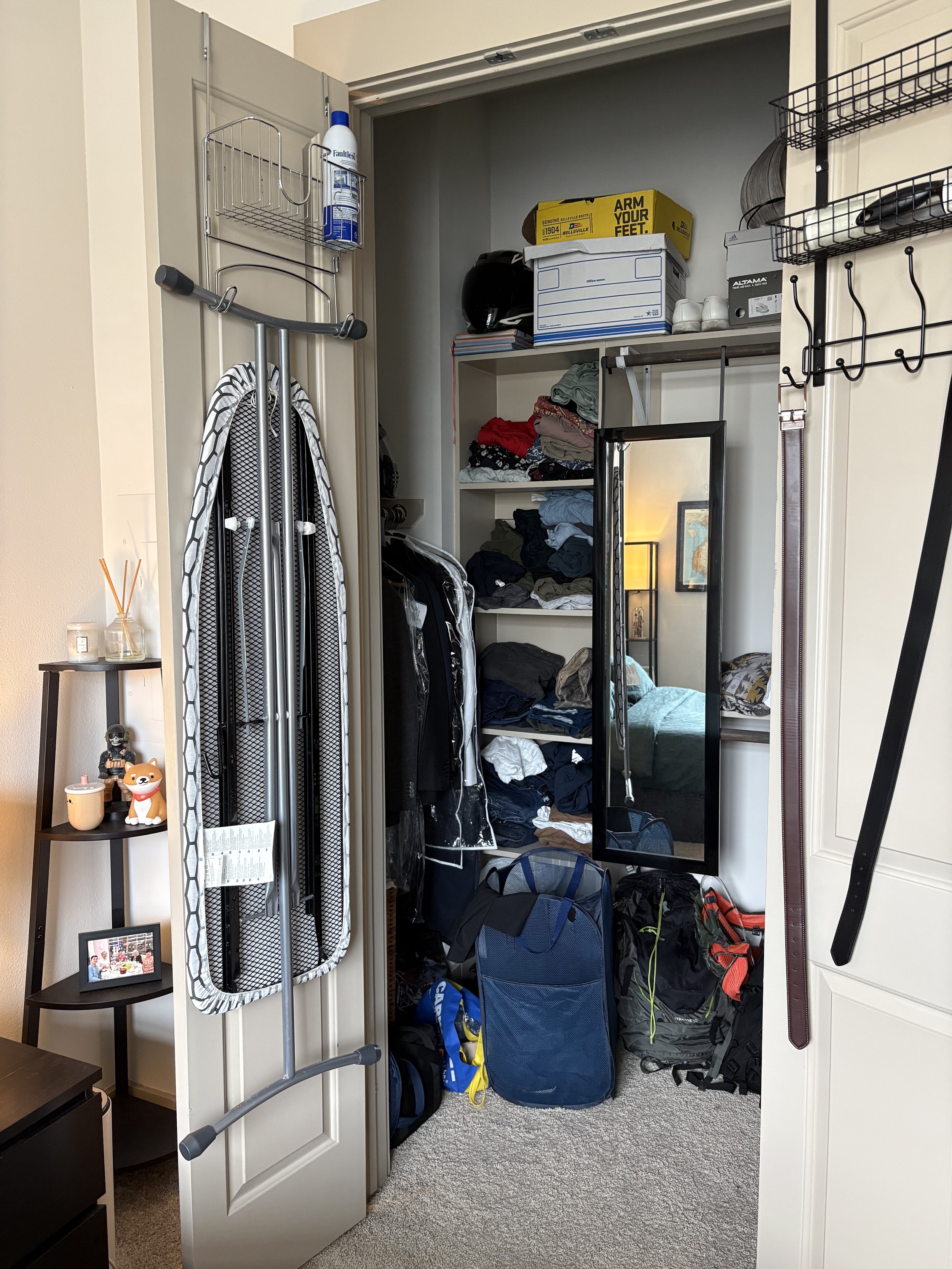 Open closet with shelves, clothes, backpacks, and a mirror, alongside a small shelf with decorative items and a dissolving reed diffuser.