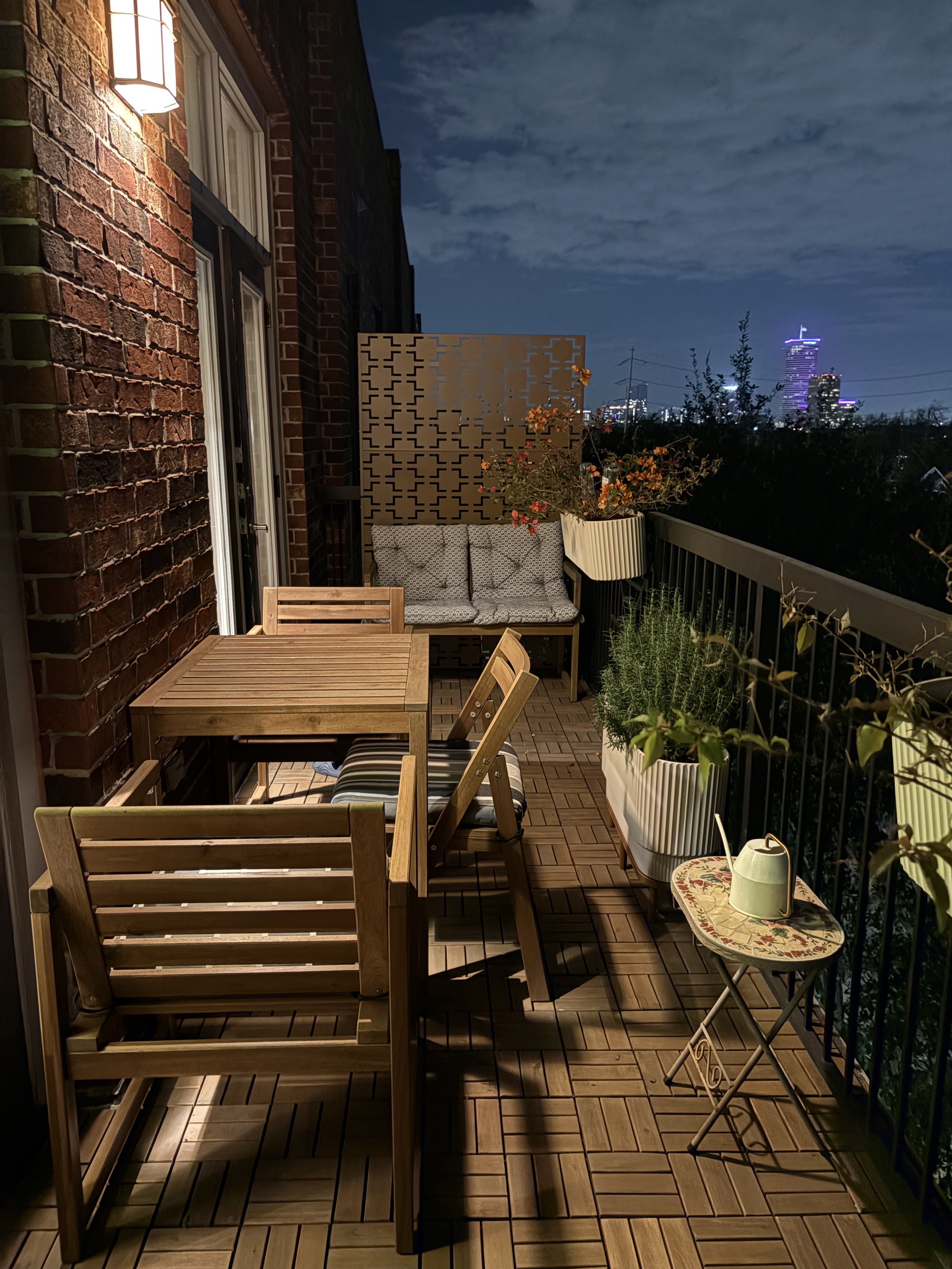 Night view of a small balcony with wooden furniture, potted plants, cushion seating, and city skyline in the background.