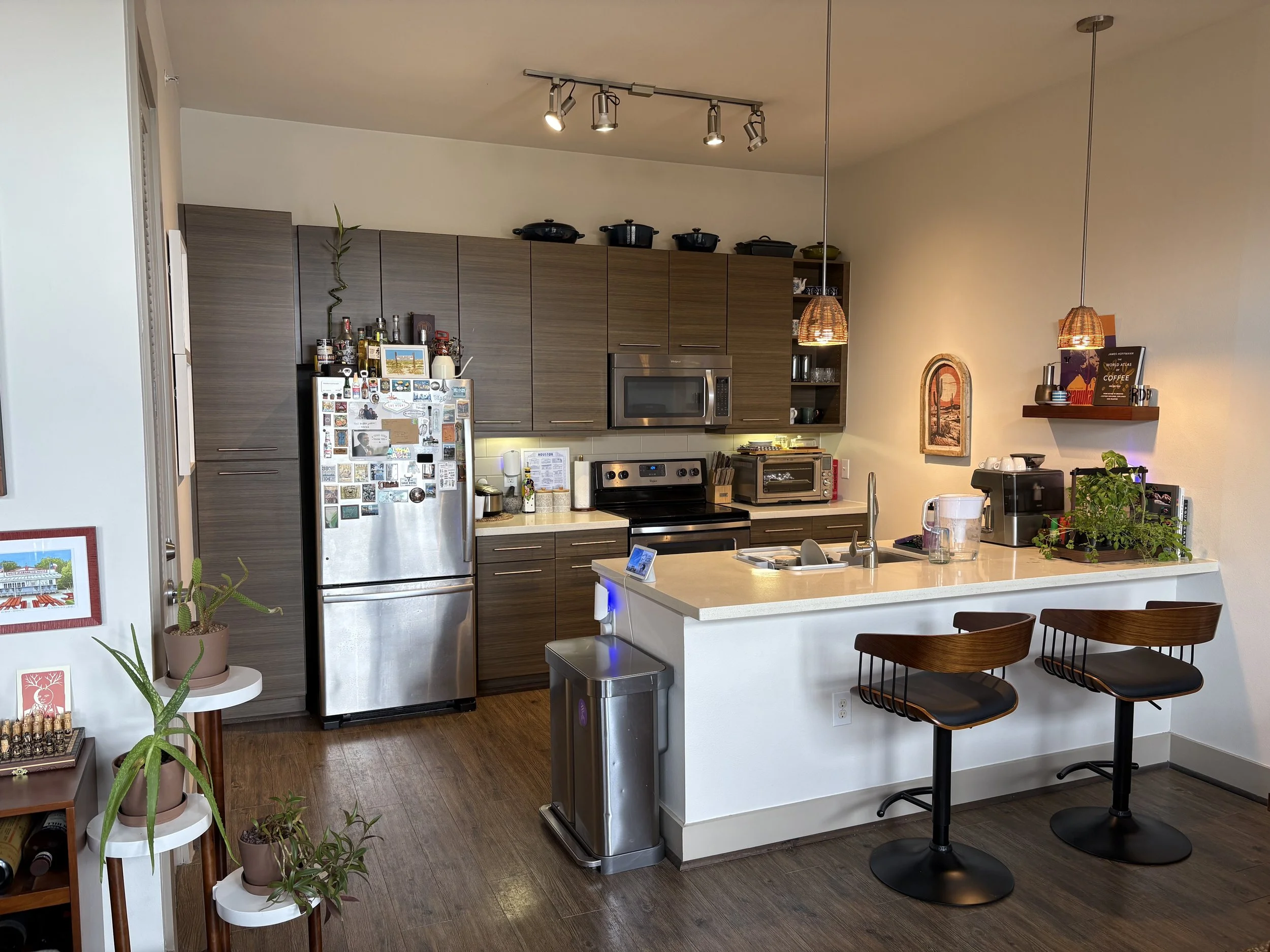 Modern kitchen with wooden cabinets, stainless steel refrigerator, microwave, toaster oven, coffee machine, plants, and bar stools at a white countertop.