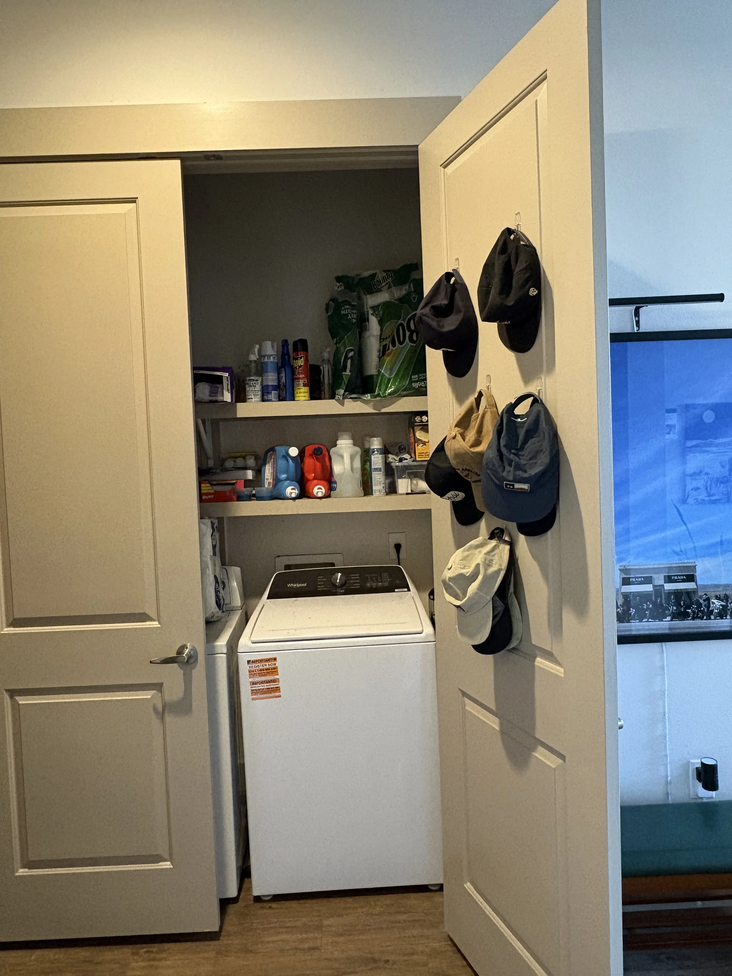 A laundry room with a washing machine, shelves with cleaning supplies, and hats hanging on the door.
