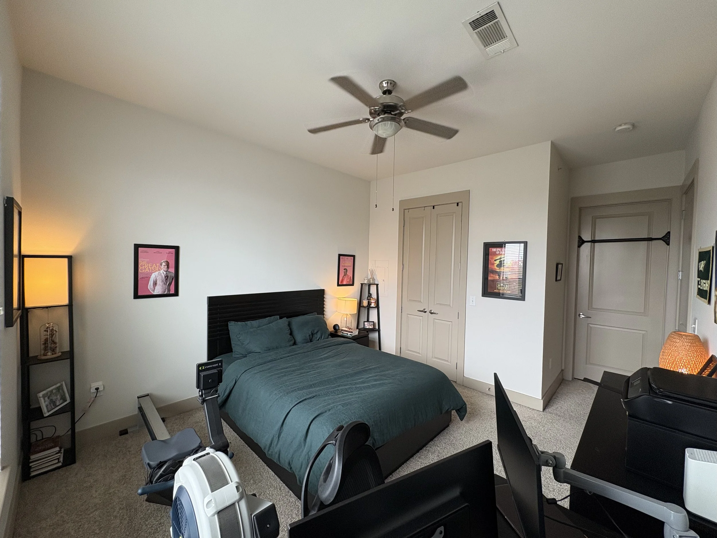 A bedroom with a bed, black headboard, teal bedding, framed art on the wall, a ceiling fan, a small lamp, a shelving unit, a closed closet with double doors, and a desk with chairs and computer monitors.