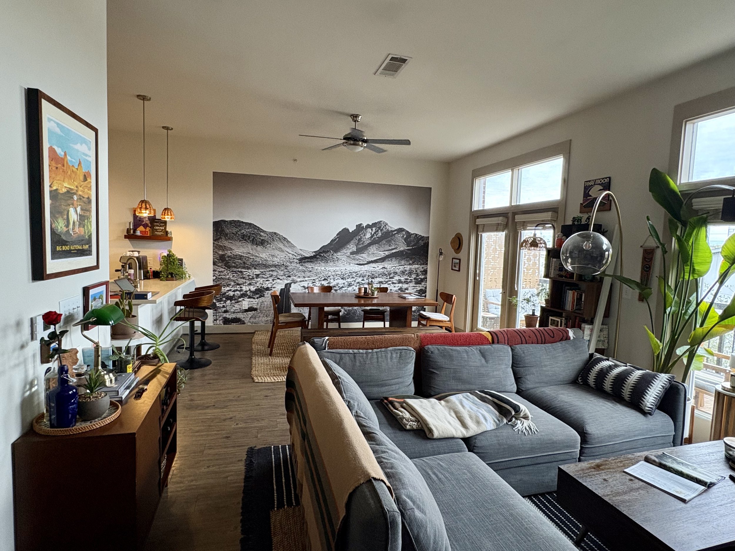 Living room with gray sectional sofa, wooden coffee table, large black and white mountain mural on the back wall, dining table with chairs, large windows, houseplants, bookshelf, and decorative artwork.