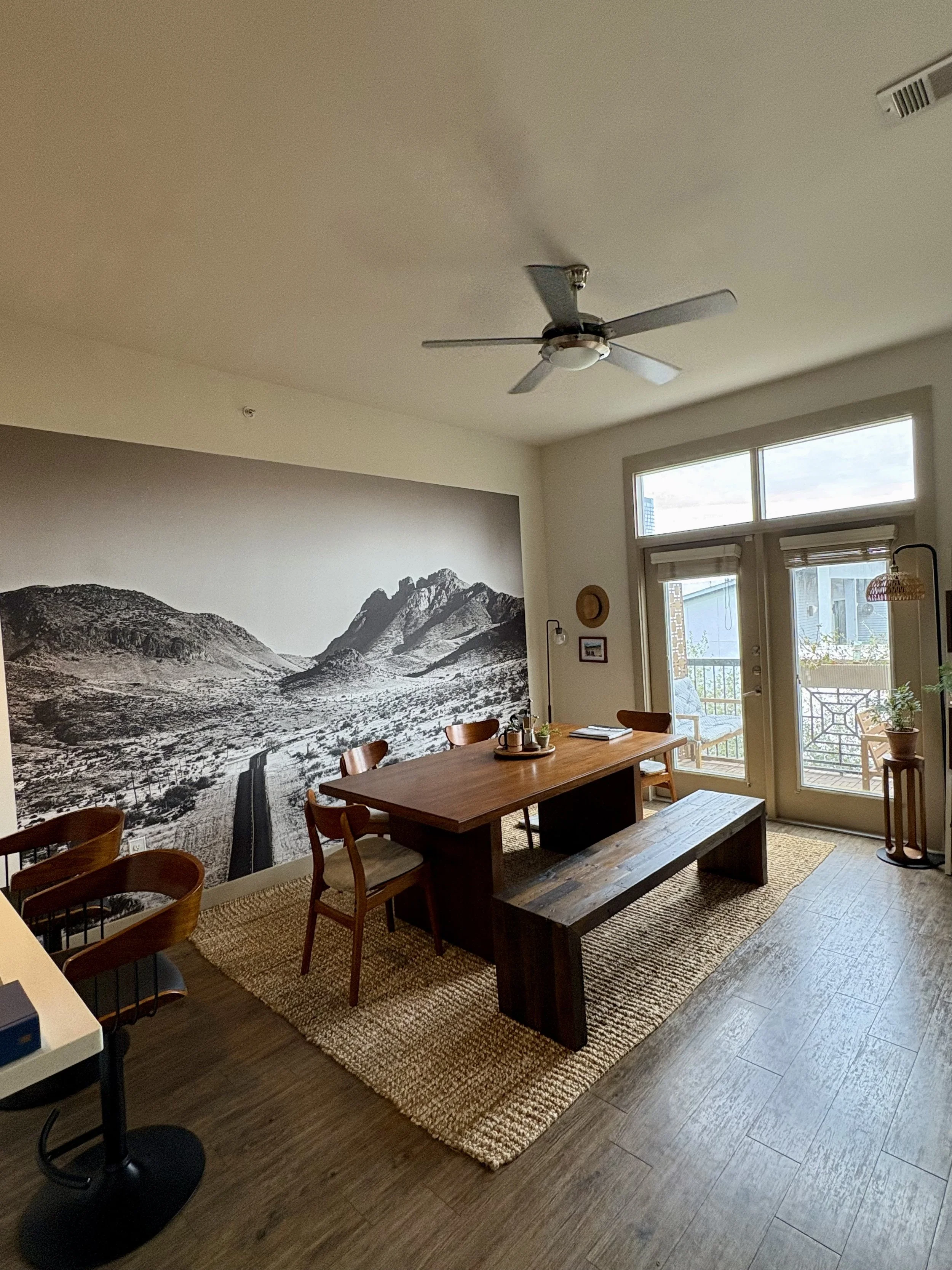 Dining room with a wooden table, chairs, a bench, a large mural of mountains on the wall, and a sliding glass door leading to a balcony.