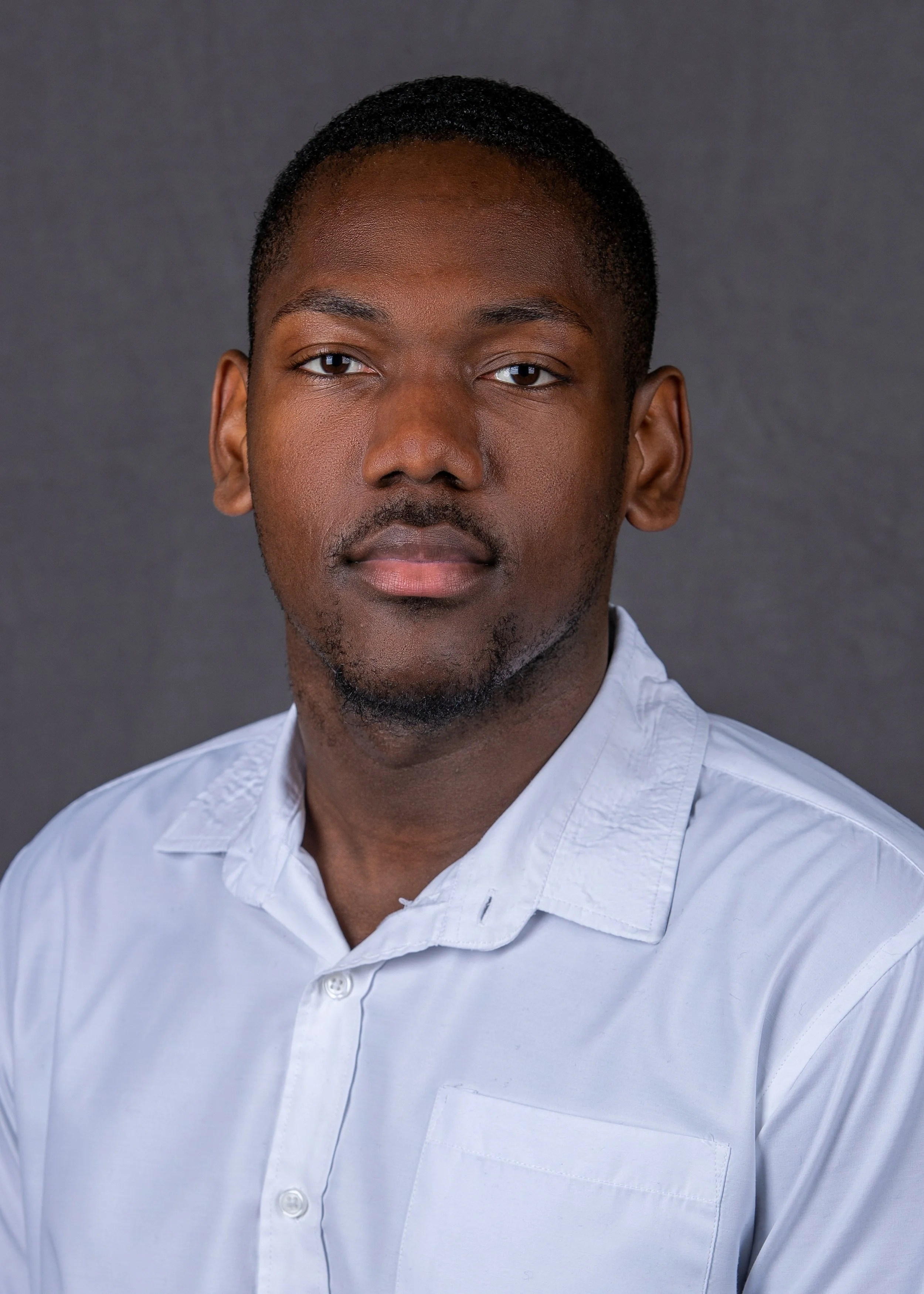 A young man with short black hair and a goatee wearing a white button-up shirt against a dark gray background.