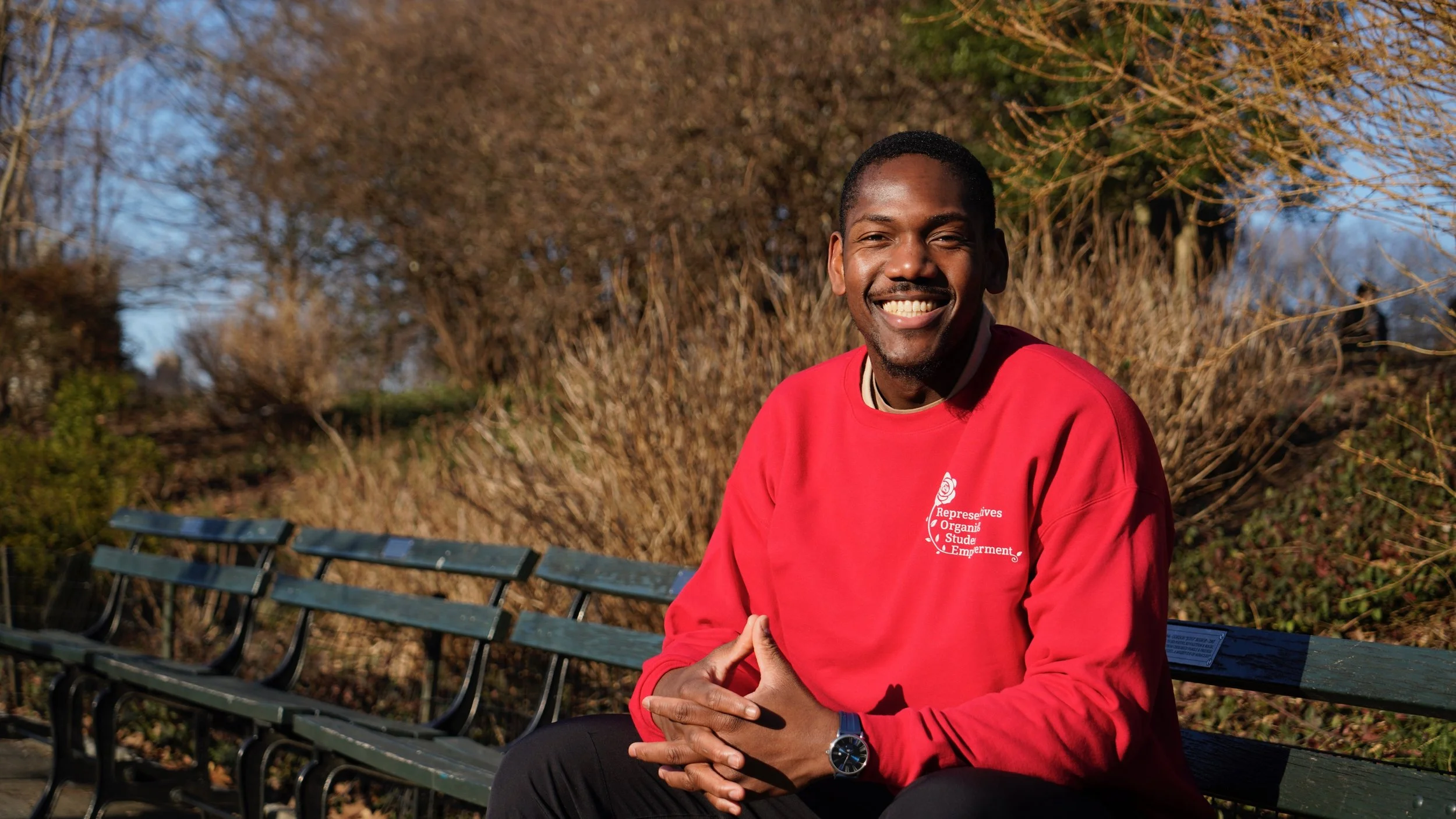 A smiling man with dark skin, wearing a red sweatshirt, sitting on a park bench with his hands clasped, in a sunny outdoor setting with trees and bushes in the background.