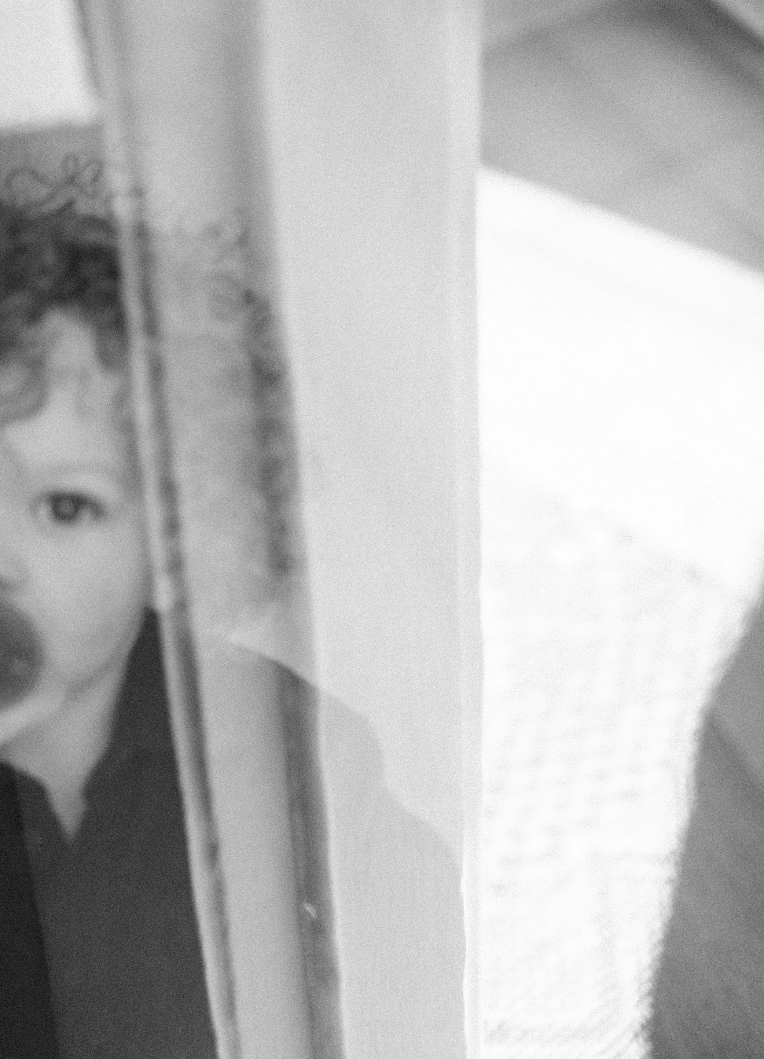 A blurry black and white photo showing part of a portrait of a woman with curly hair, partially obscured by a vertical frame, next to a bright window with blinds.