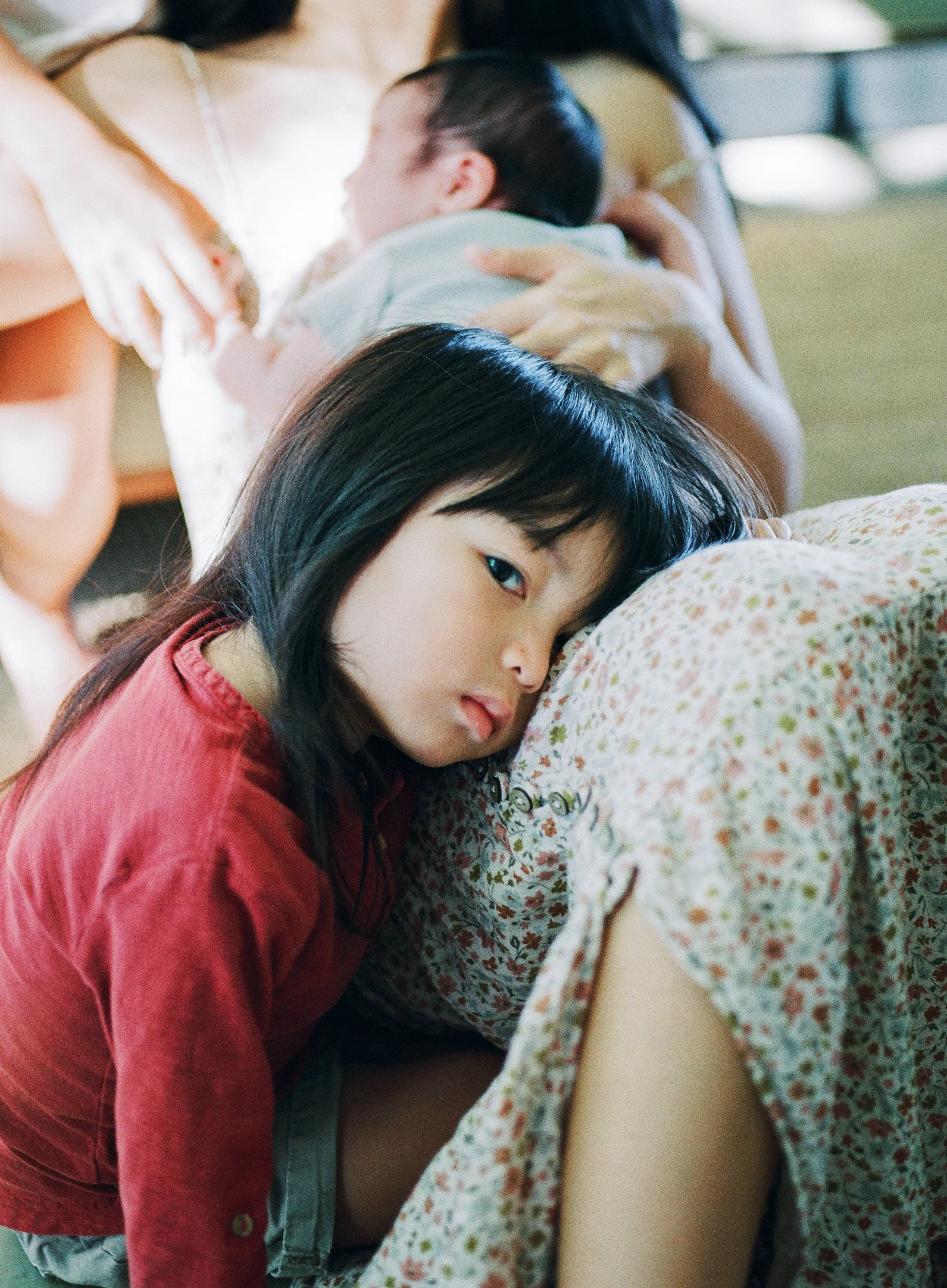 A young girl rests her head on a floral pillow, looking into the camera with a calm expression, while a woman in the background is holding an infant.