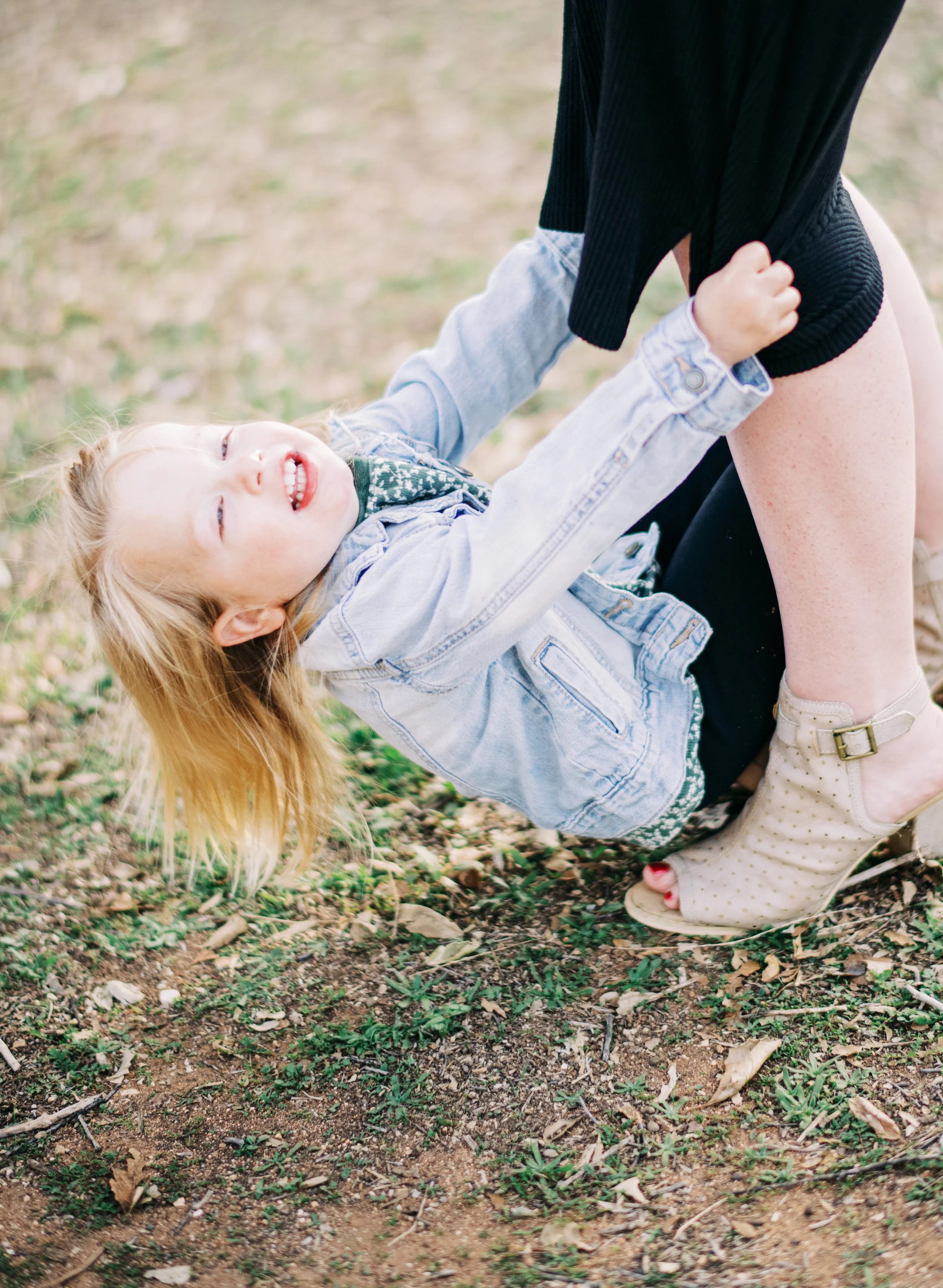 A young girl with blonde hair and a denim jacket on her shoulders, smiling and looking joyful, is playing on the ground outdoors with an adult, possibly her mother. The girl is lying backwards on the soil, holding onto the adult's knees.