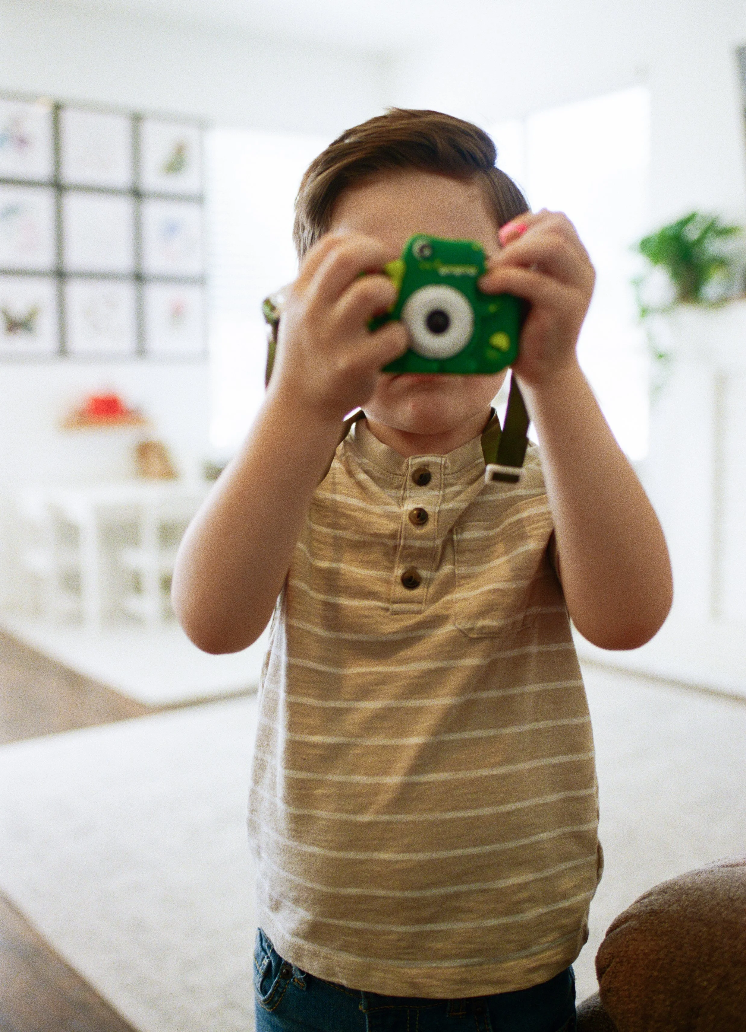 A young boy in a beige striped shirt holds a green toy camera up to his face, taking a photo.