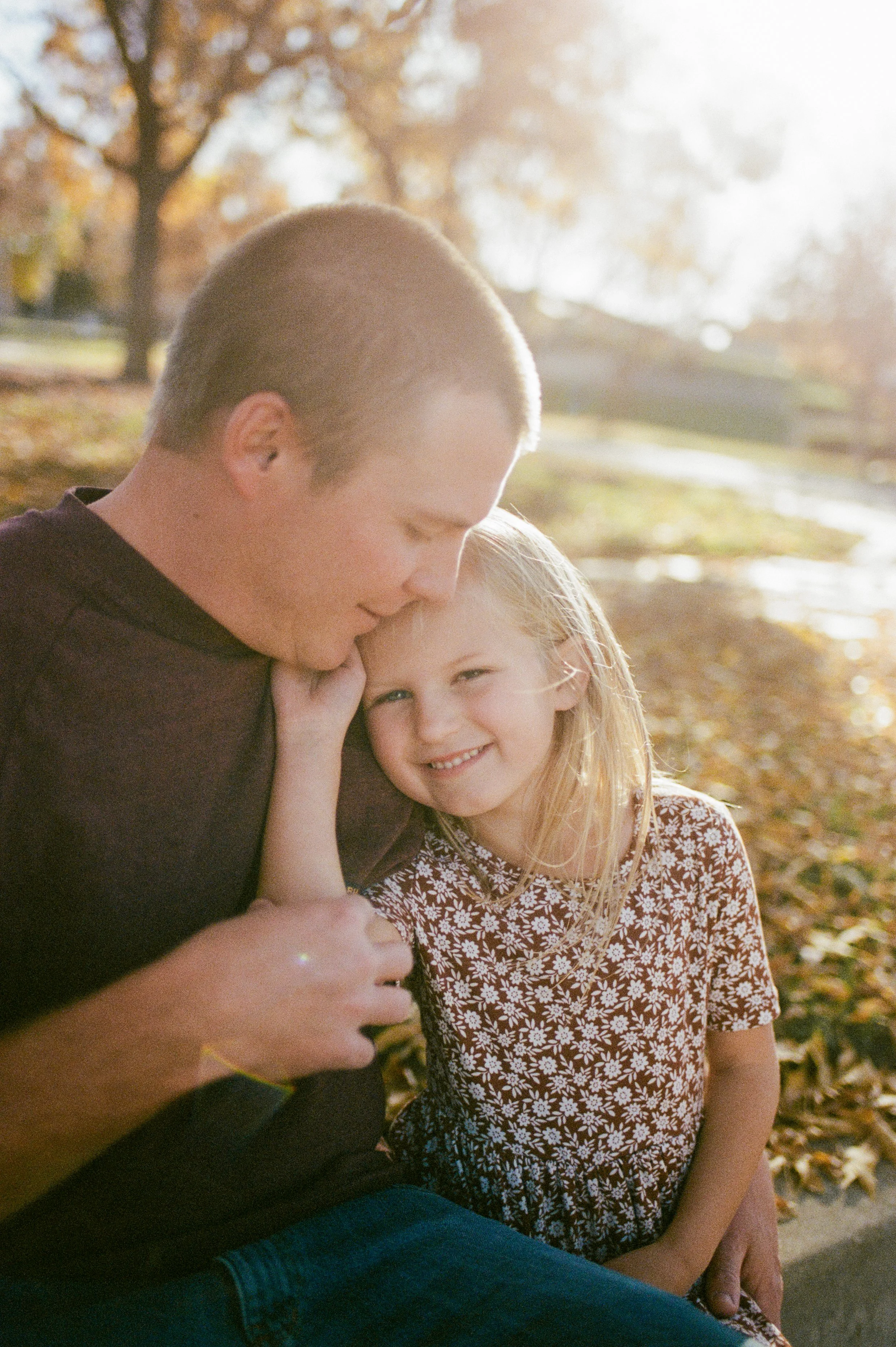 A man and a young girl sitting outdoors on fallen autumn leaves, smiling and enjoying each other's company in the sunlight.