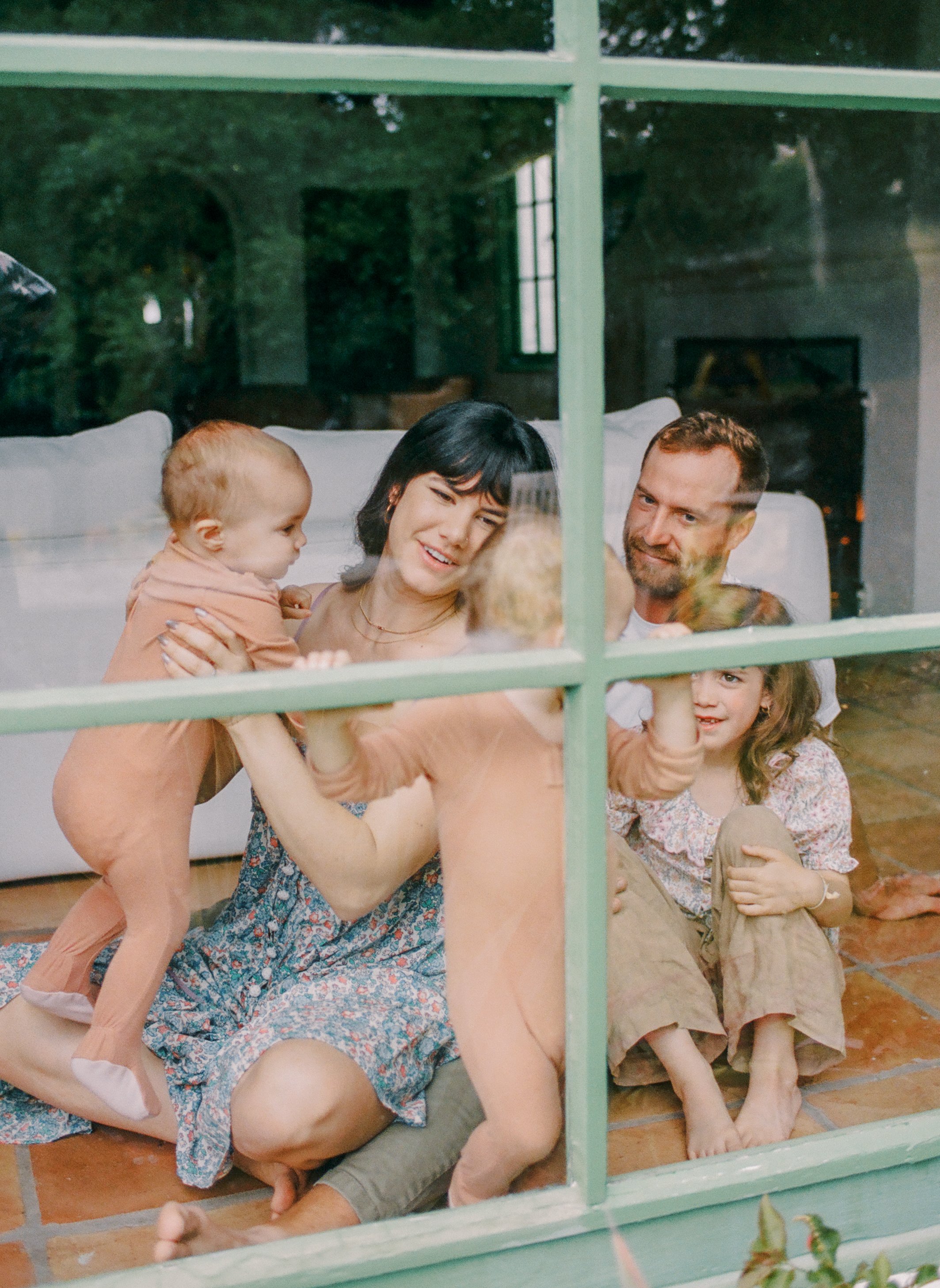 A family of four seen through a glass window, with children playing on the floor and the parents smiling and interacting inside a cozy living room.
