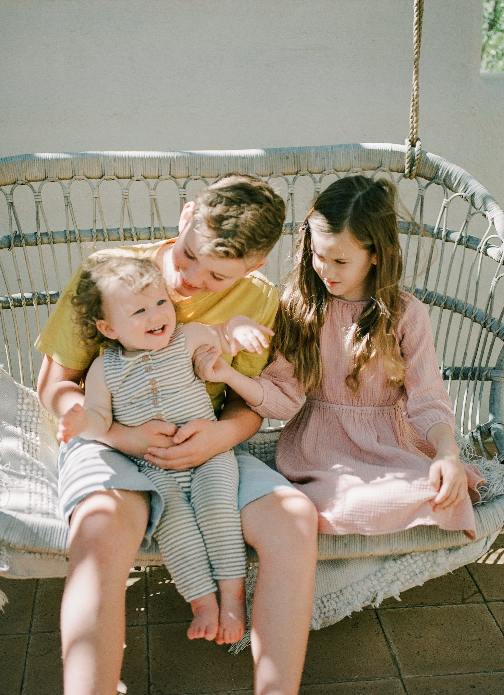Three children sitting on a hanging wicker chair, with a toddler girl laughing and two older children smiling and interacting with her, outdoors in sunlight.