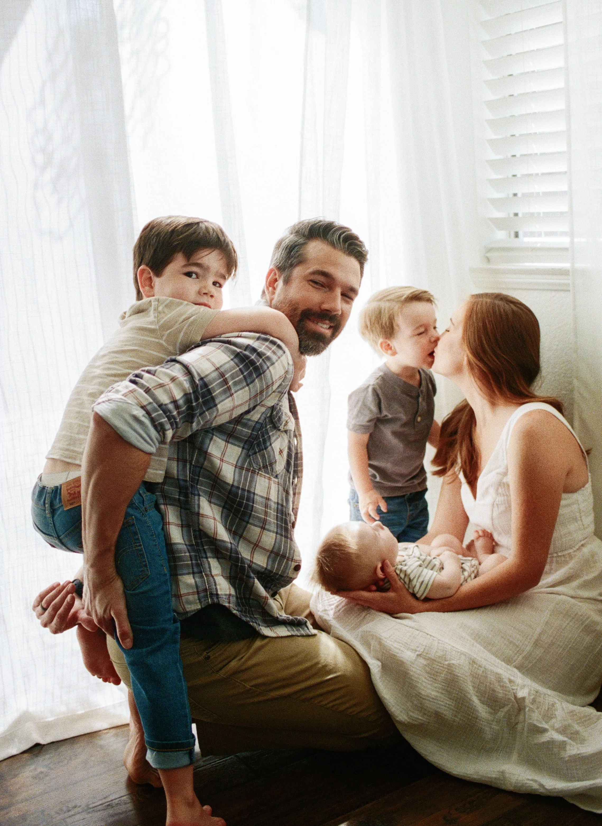 A family of five in a bright room, with a woman sitting on the floor holding a newborn baby, a man and two young boys nearby, one boy on the man's back and the other kissing the woman, all sharing a joyful moment.