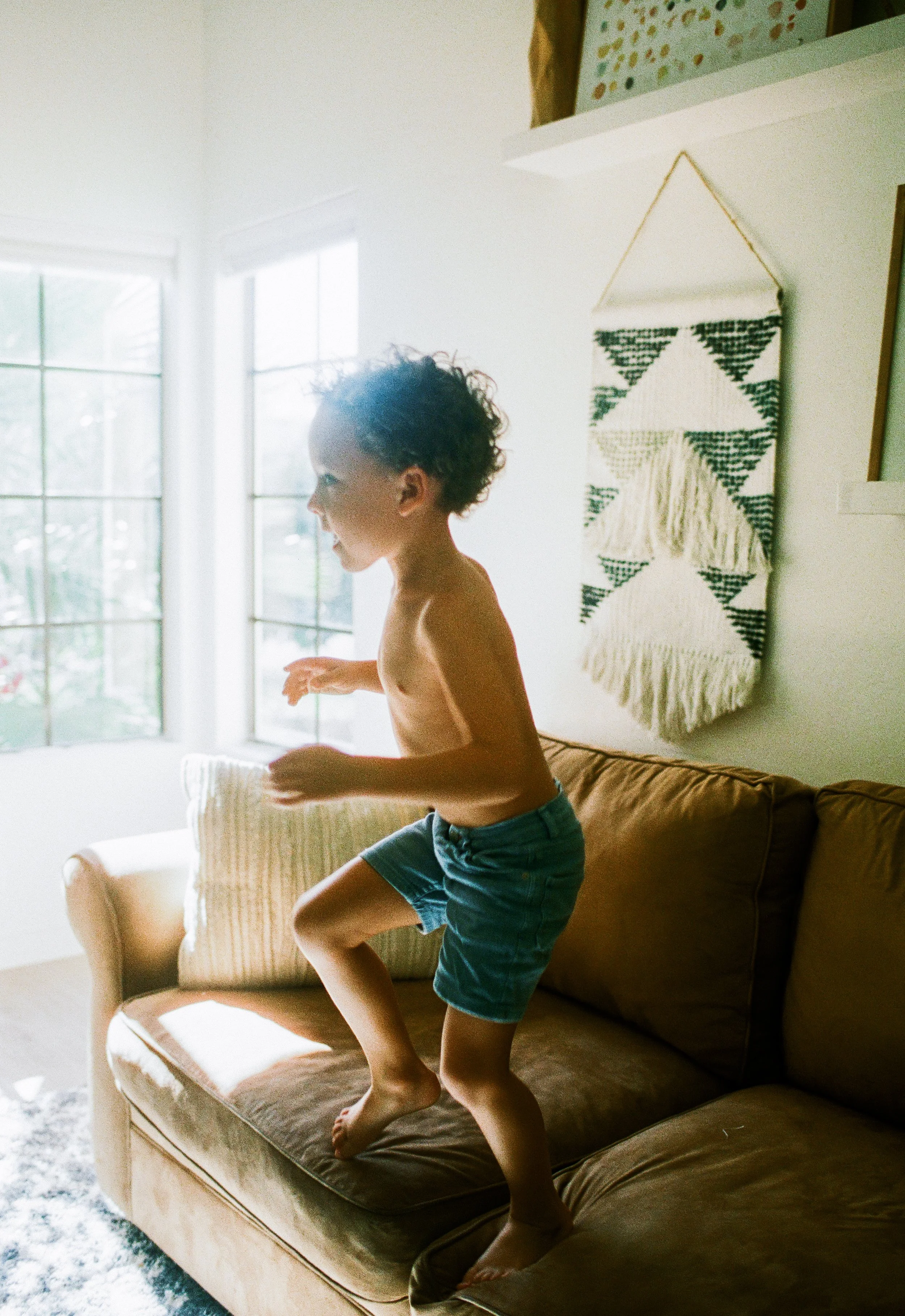 A young boy with curly hair jumping on a brown couch in a sunlit living room