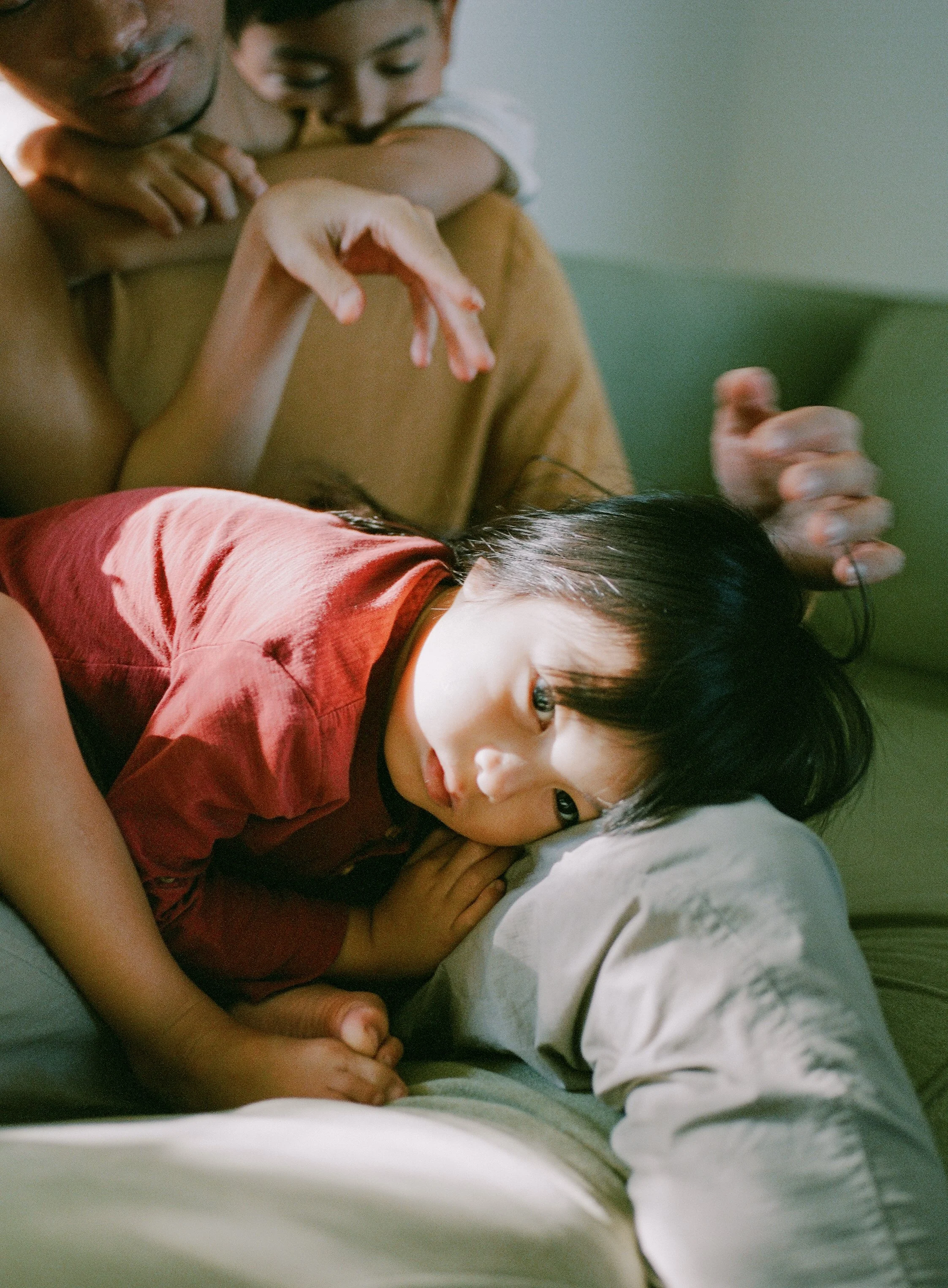 A young girl lying on her side on a bed, resting her head on a pillow, with a somber expression, while adults surround her, with one adult holding her arm and another adult reaching over her.