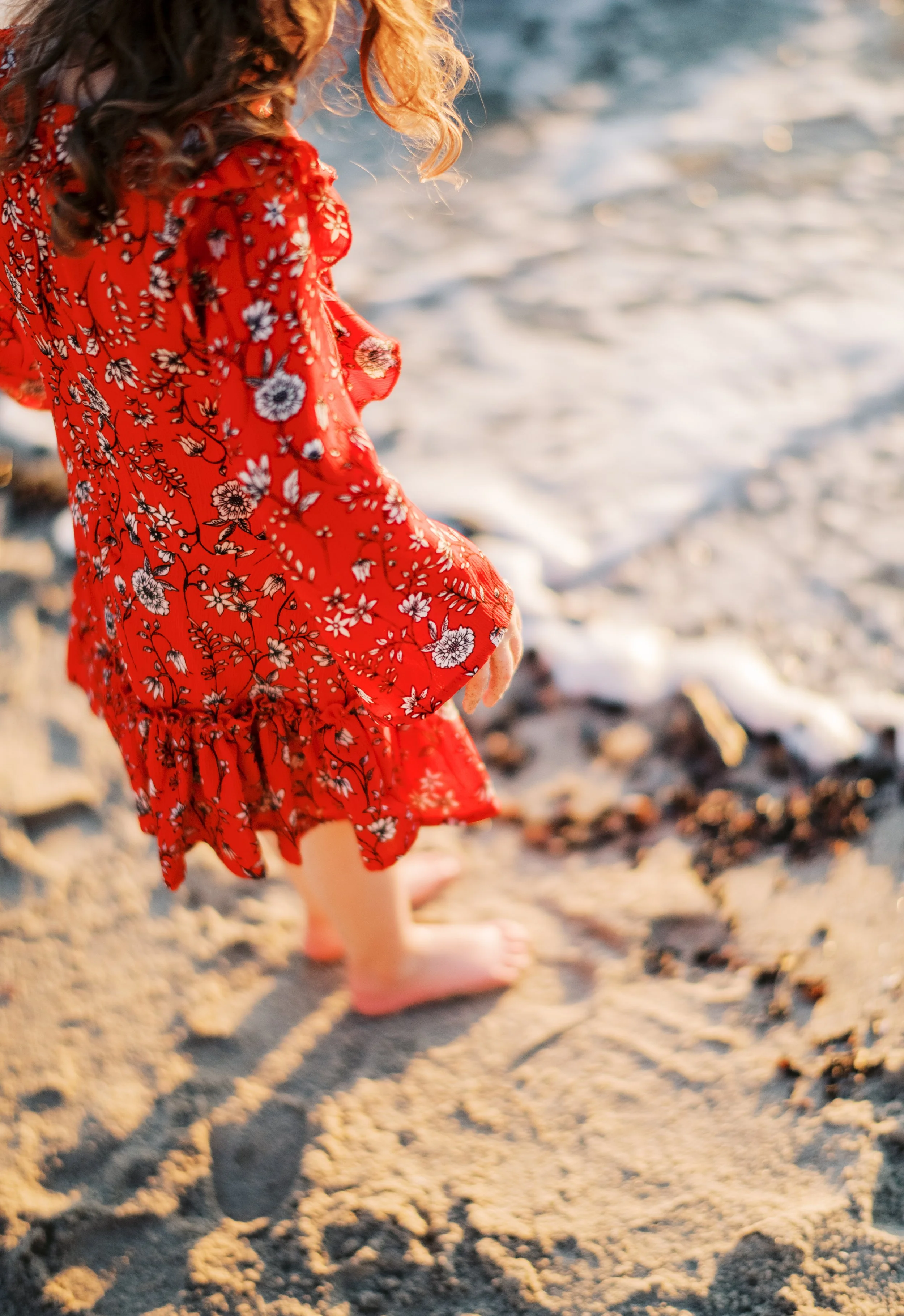 A young girl in a red floral dress standing barefoot on a sandy beach near the water, with her feet partially in the ocean waves.