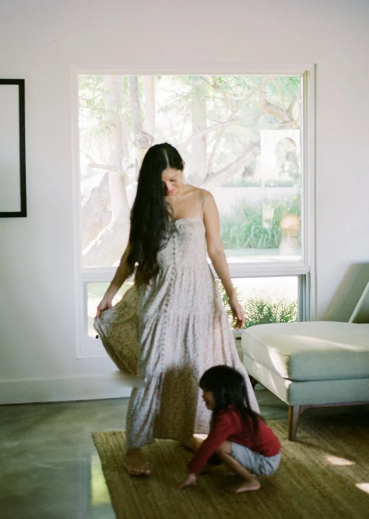 A woman with long dark hair wearing a light-colored, patterned dress standing on a brown rug, holding the hem of her dress. A young girl with dark hair wearing a red top and gray bottoms is crouching near her feet inside a room with a large window sh