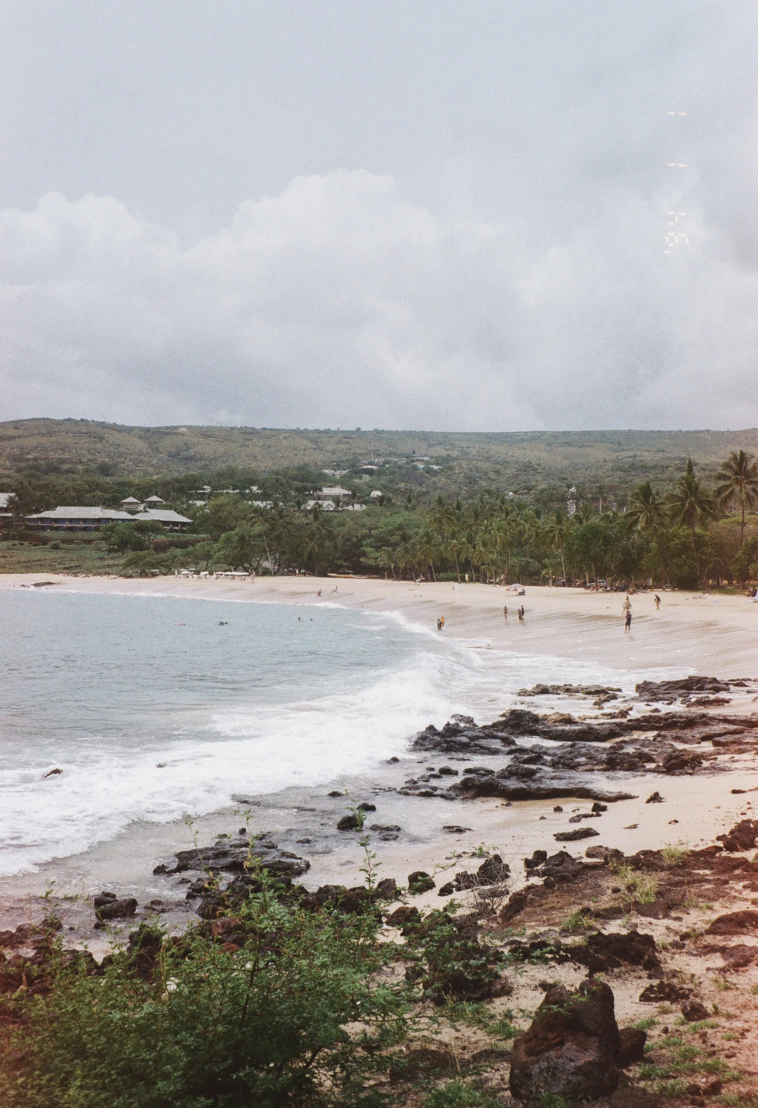 A beach with waves crashing on rocks and sand, few people walking along the shoreline, lush green trees, and buildings with a hill in the background on a cloudy day.