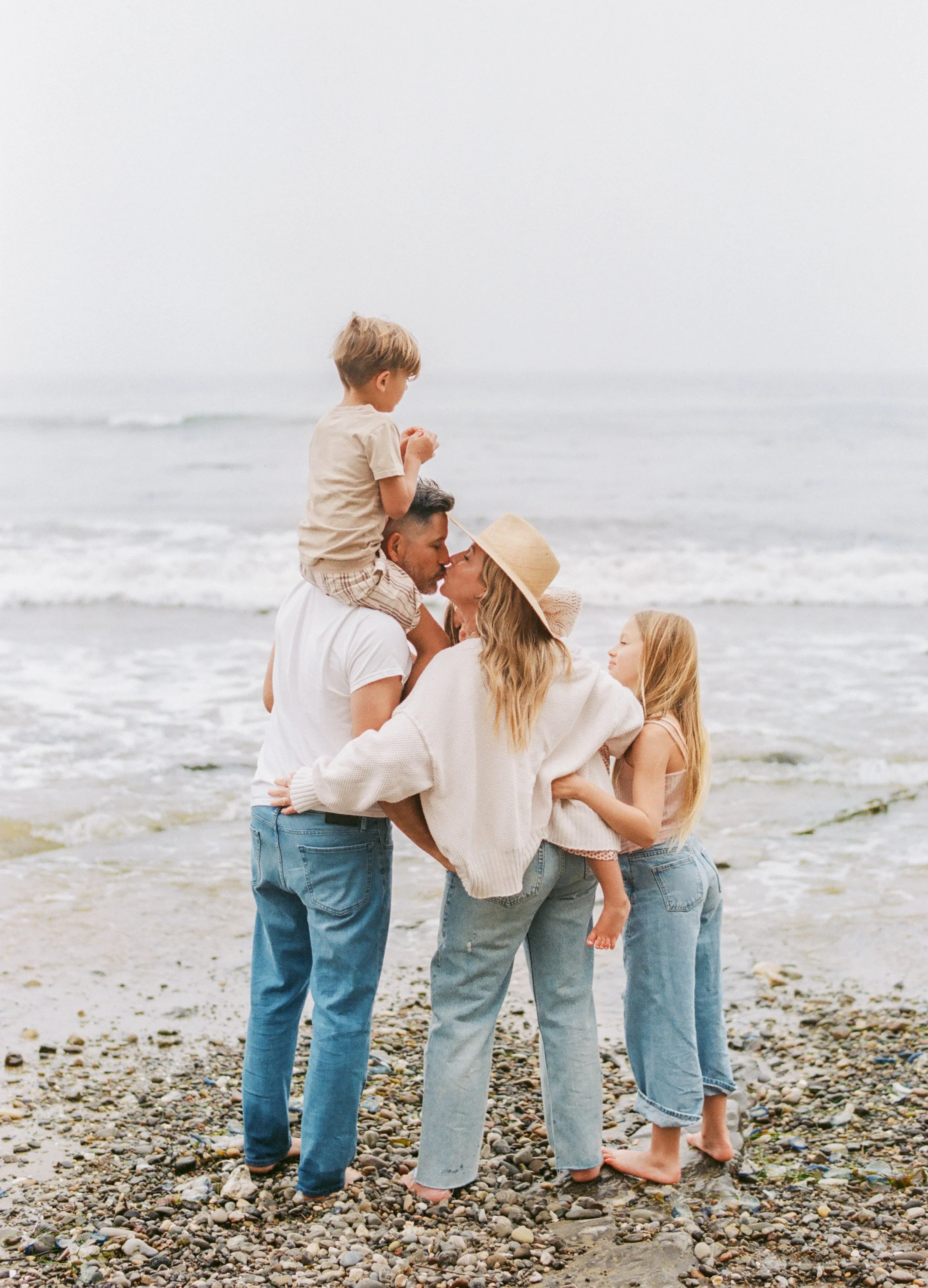A family of five on a beach, with a man, woman, and three children, engaging in a group kiss, all dressed casually, with the ocean in the background.