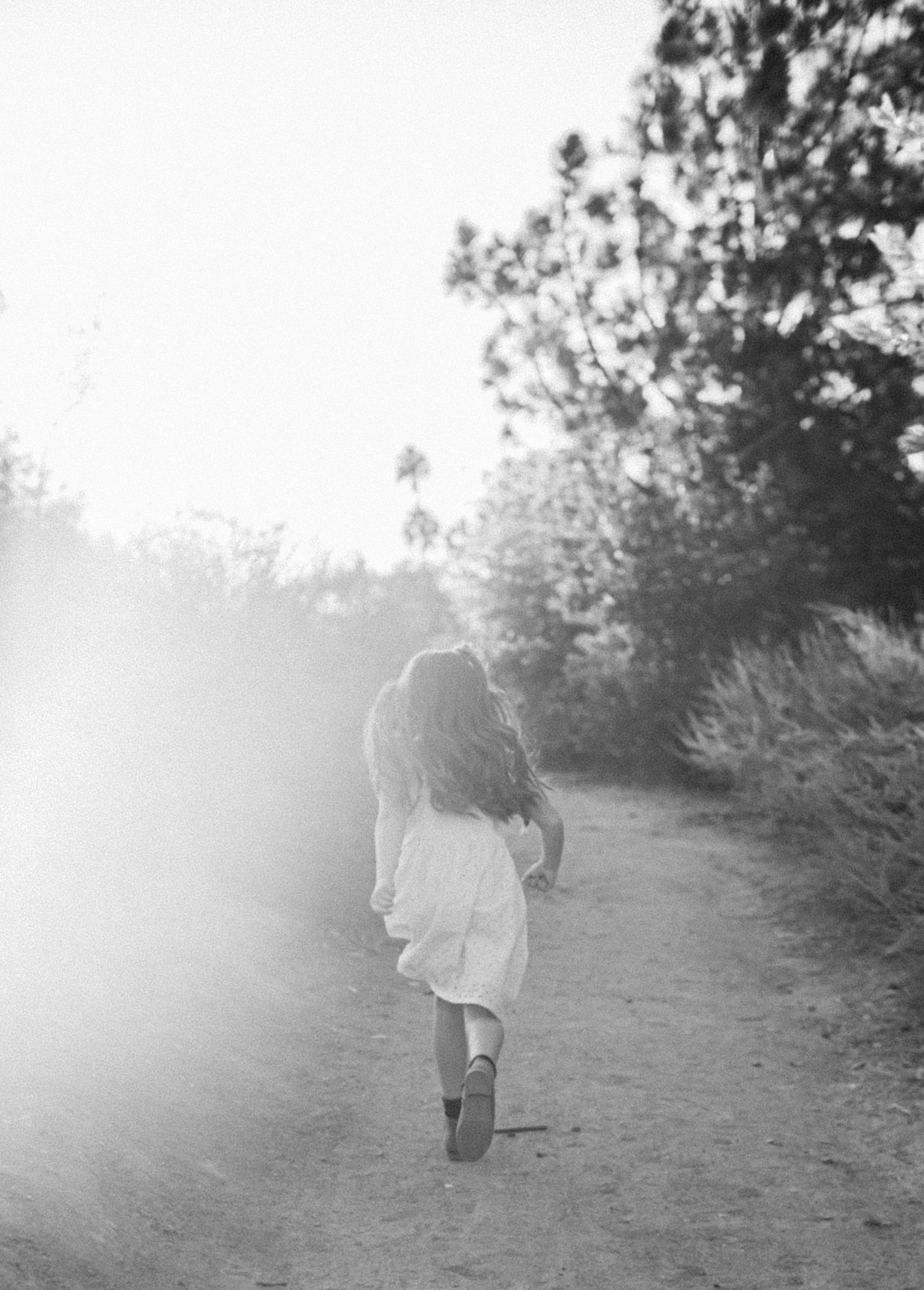 A young girl with long hair running away down a dirt path surrounded by trees, in black and white.