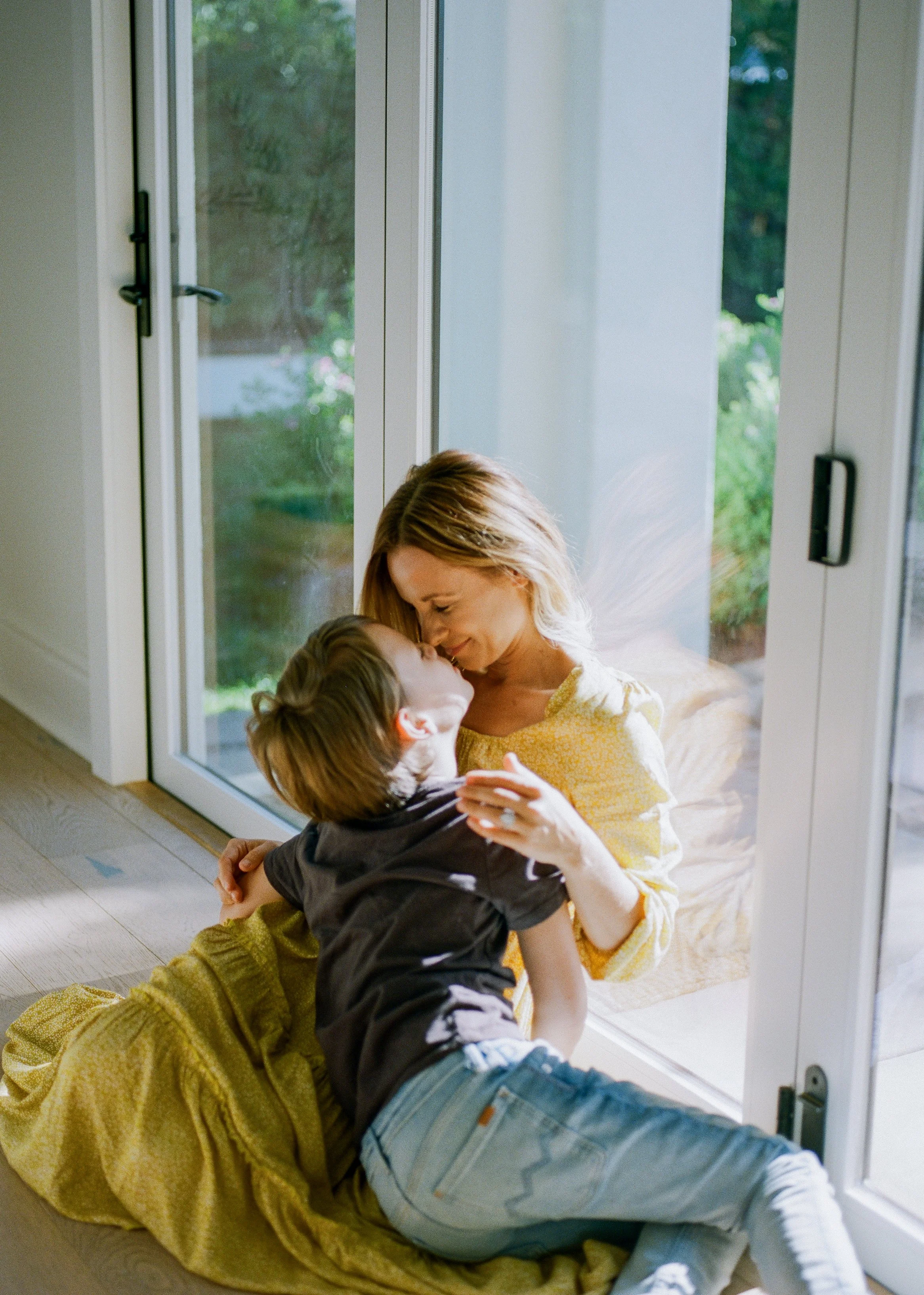 A woman and a young boy sharing a tender moment through a glass door, with the woman smiling and the boy leaning in for a kiss.