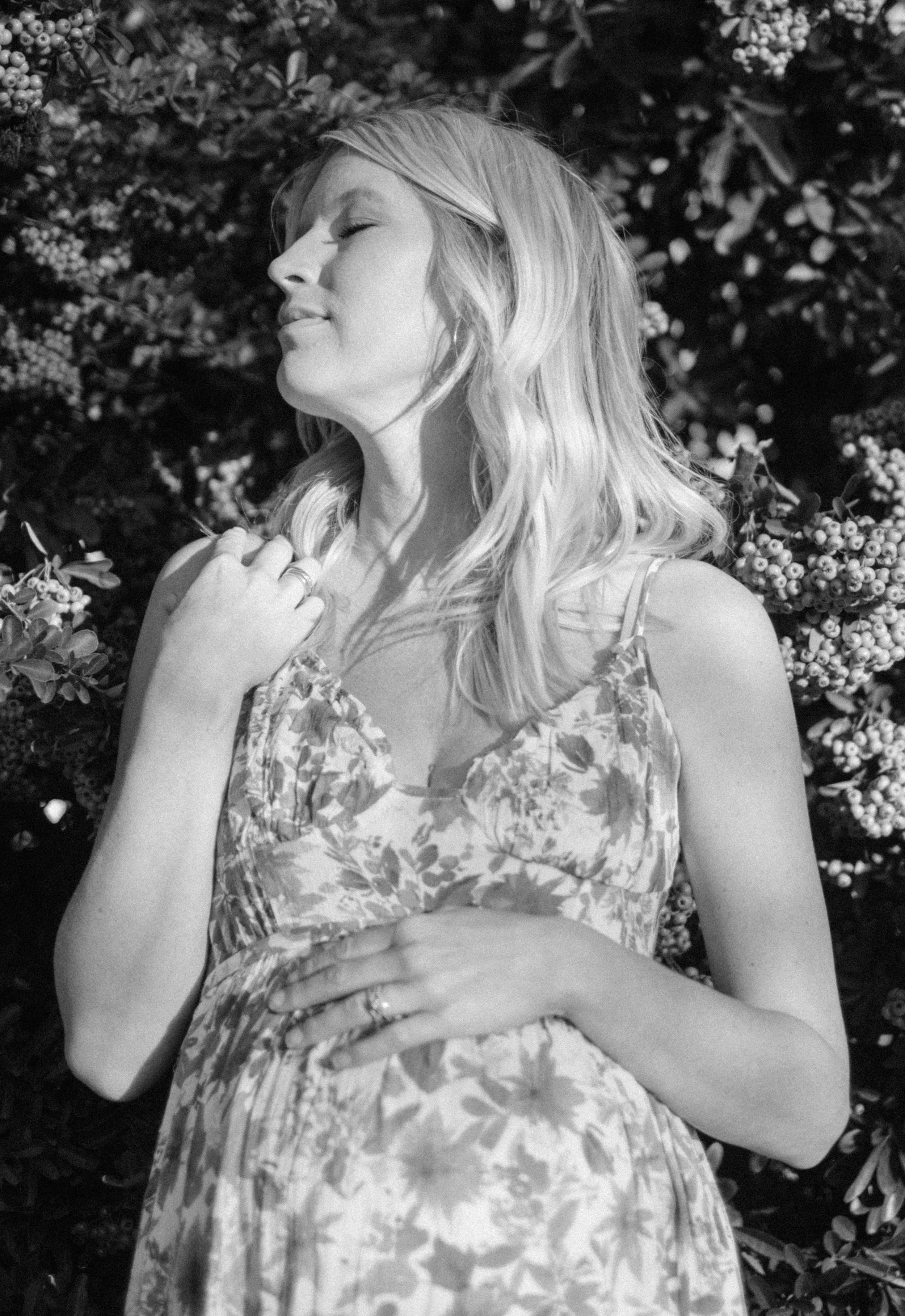A woman with long, wavy hair, wearing a floral dress, standing outdoors in front of a bush with small berries, her eyes closed and a serene expression.