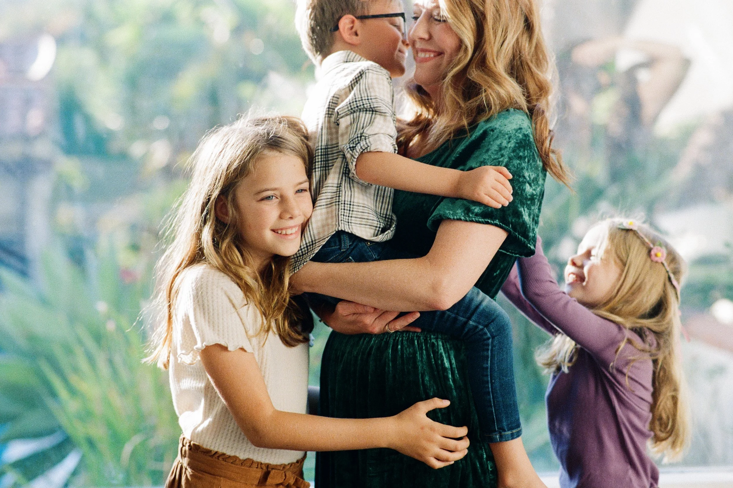 A woman is holding two children, a boy and a girl, while another girl hugs her. The woman has red hair and is smiling. The children are smiling and appear happy in an indoor setting with natural light.