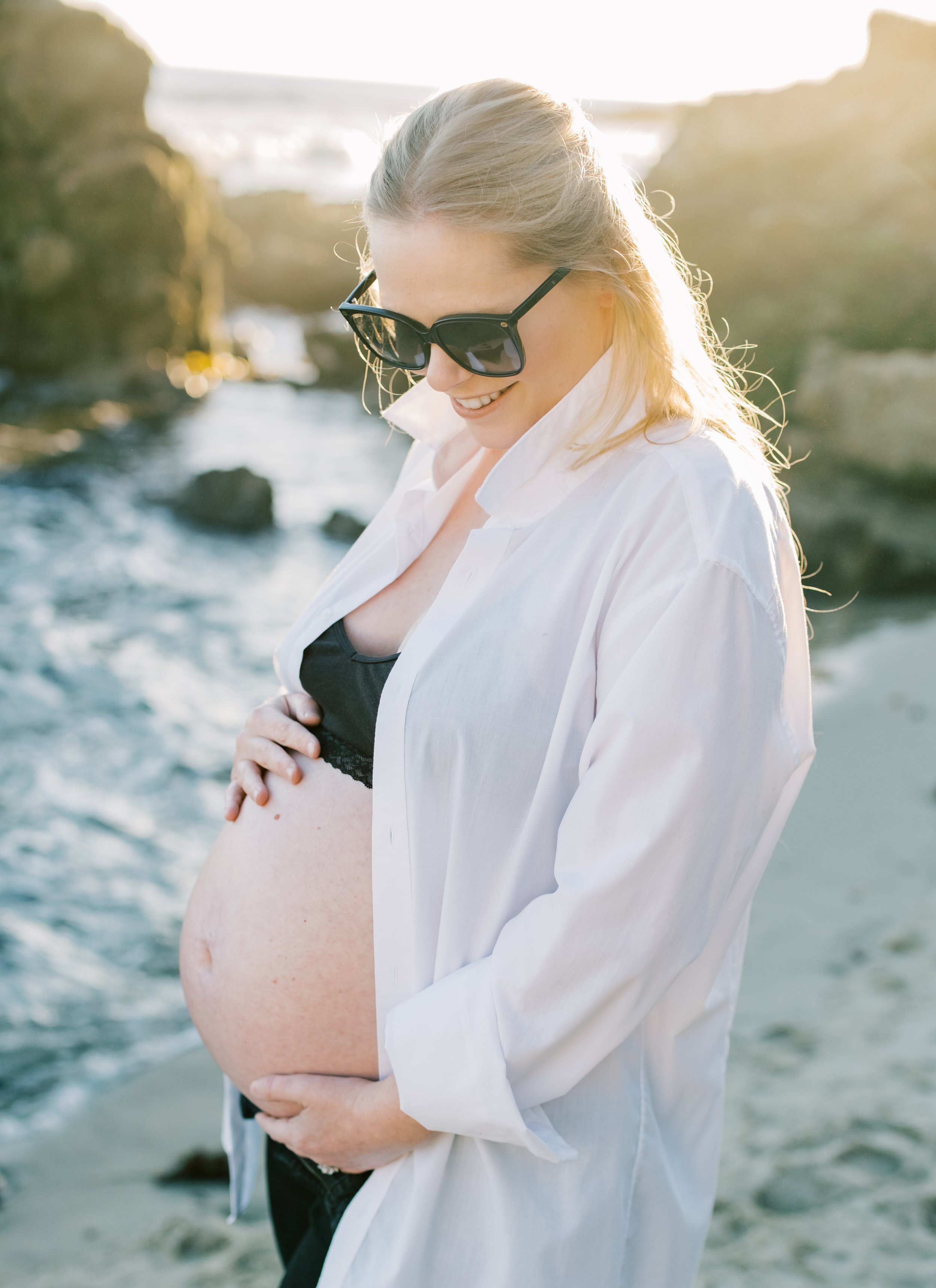 A pregnant woman in sunglasses and a white shirt standing on a beach, smiling and touching her belly with one hand, with water and rocks in the background during sunset.