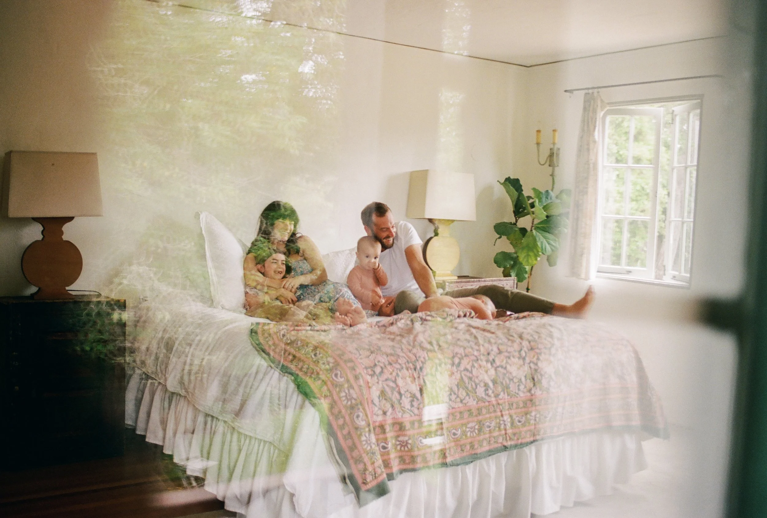 A family of four sitting on a bed in a bright room, seen through a glass window. They are smiling and laughing, with two children, a man and a woman. The room has a large window with open curtains, a bedside table with a lamp, and a potted plant.