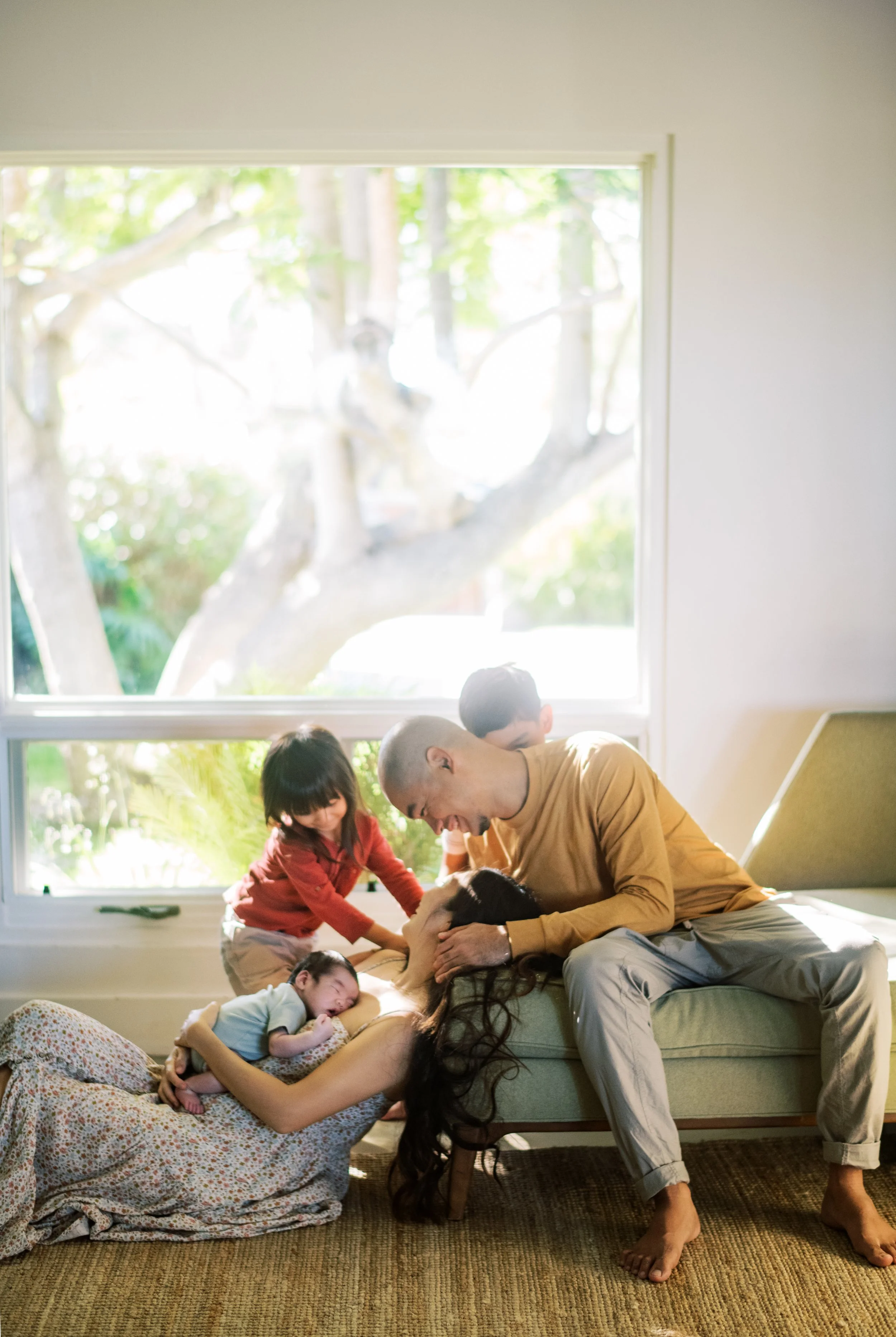 Family enjoying a moment together in a bright living room, with a large window showing trees outside, including a tree with a birdhouse.