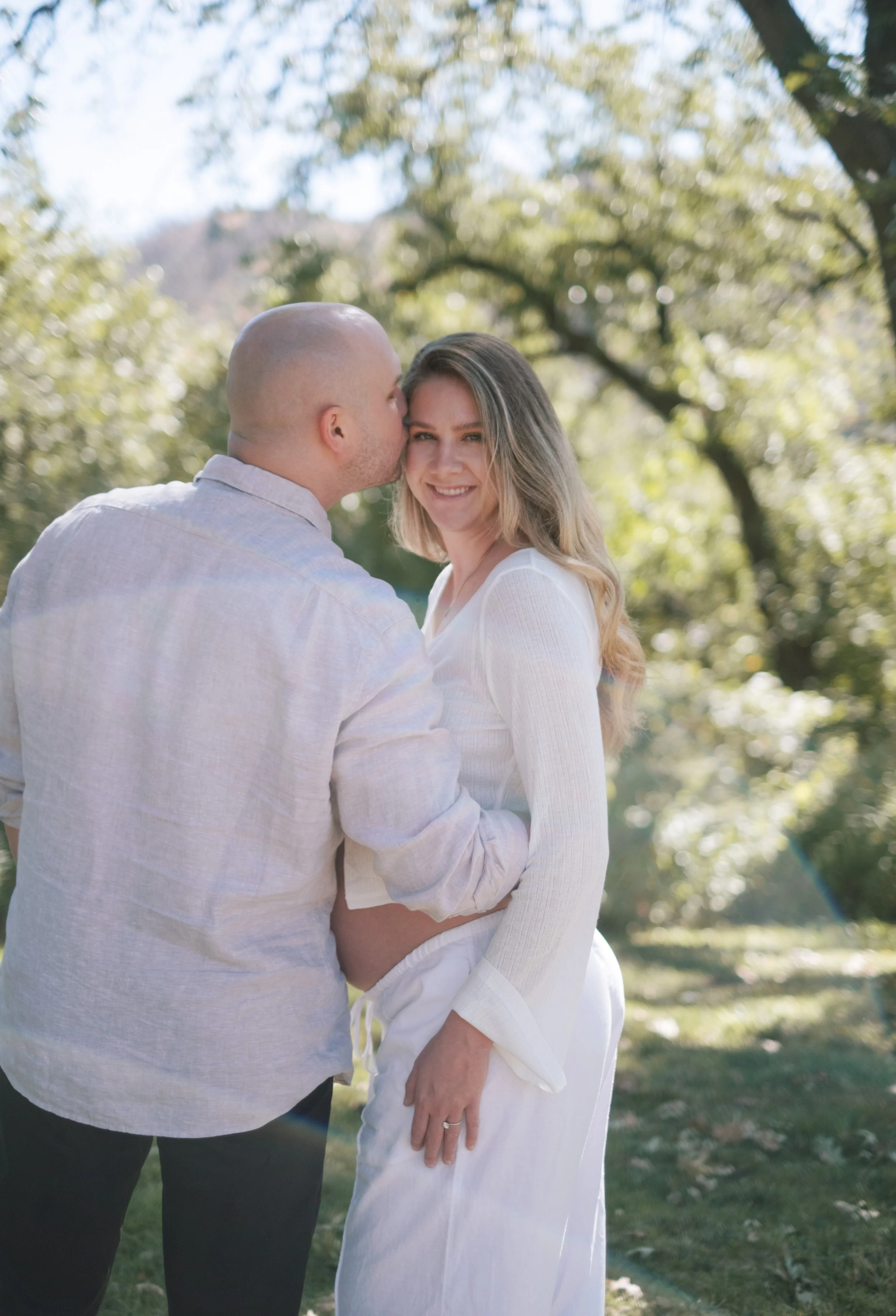 A couple standing outdoors, with the man kissing the woman on the cheek. The woman is smiling and looking at the camera, with trees and sunlight in the background.
