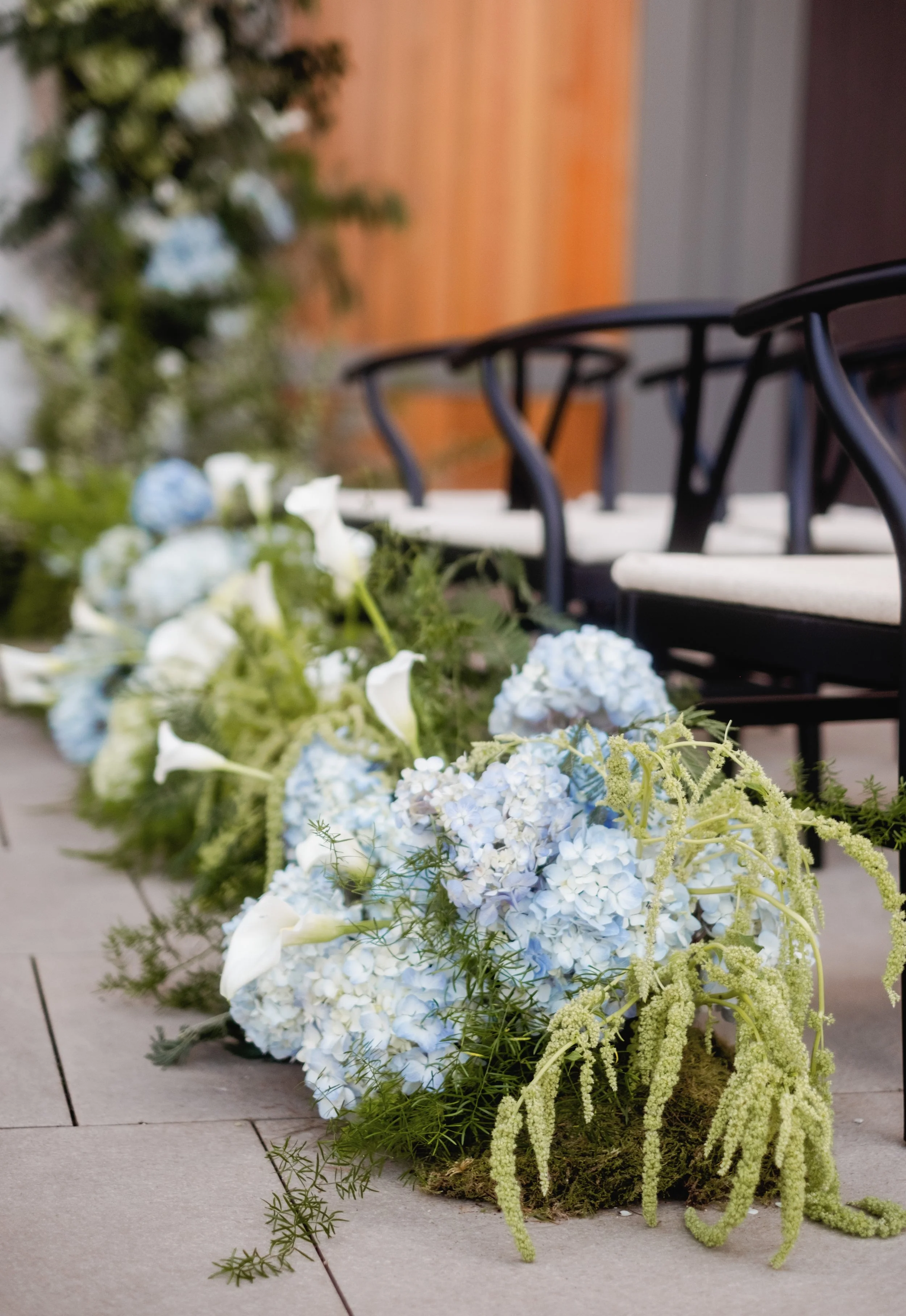 A floral arrangement with white and light blue hydrangeas, white calla lilies, and green foliage placed on the ground beside outdoor chairs with black frames and white cushions.