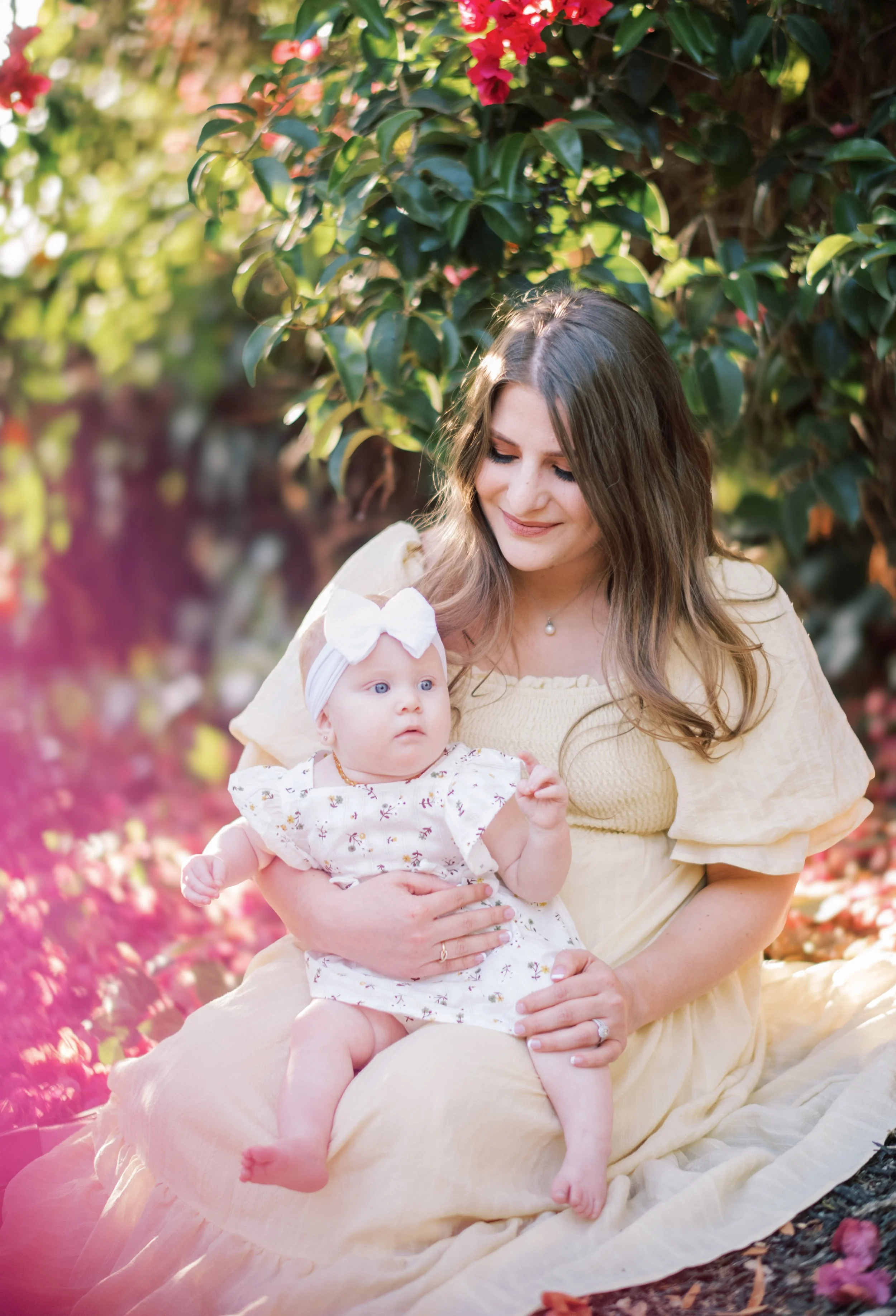 A woman with long brown hair holding a baby girl with a white headband and floral dress, sitting on pink flower petals outdoors with green foliage in background.