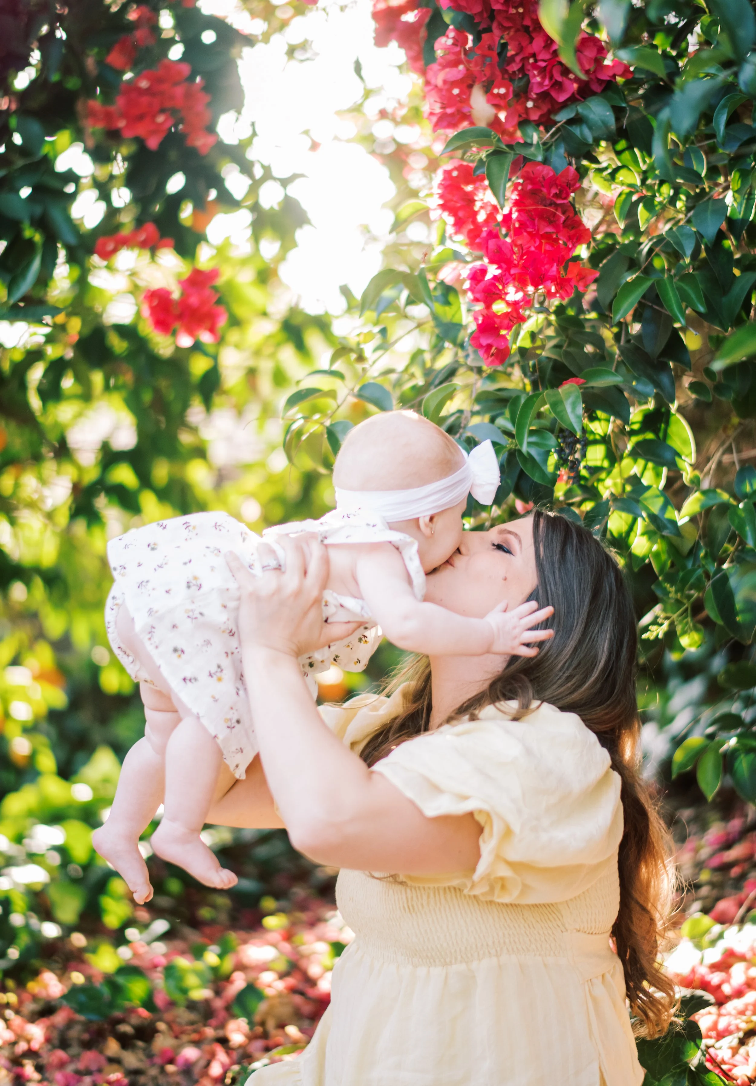 A woman holding a baby and kissing them in a garden with pink and red flowers and green foliage, with sunlight filtering through the leaves.