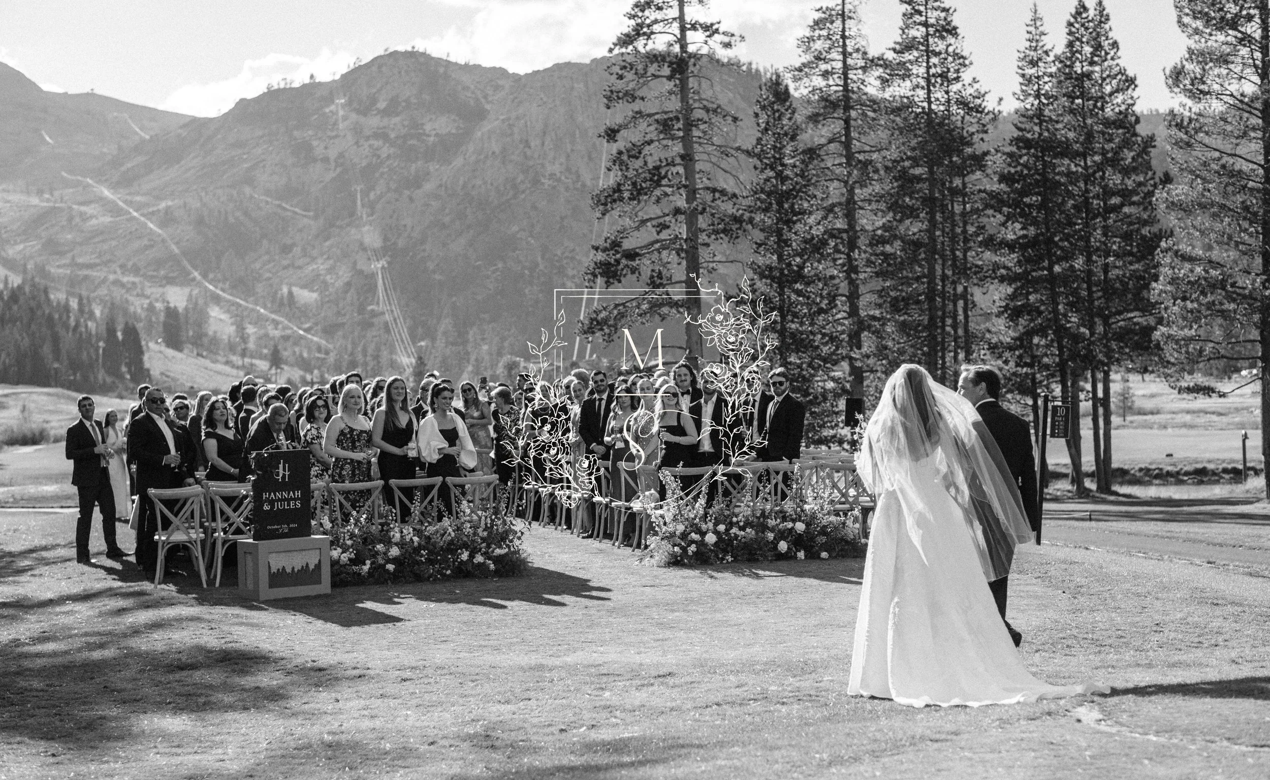 A black and white photo of a wedding ceremony outdoors with mountains and pine trees in the background. The bride and groom are facing each other, with the bride wearing a long wedding gown with a veil. Guests are standing on either side of the aisle, watching the ceremony.