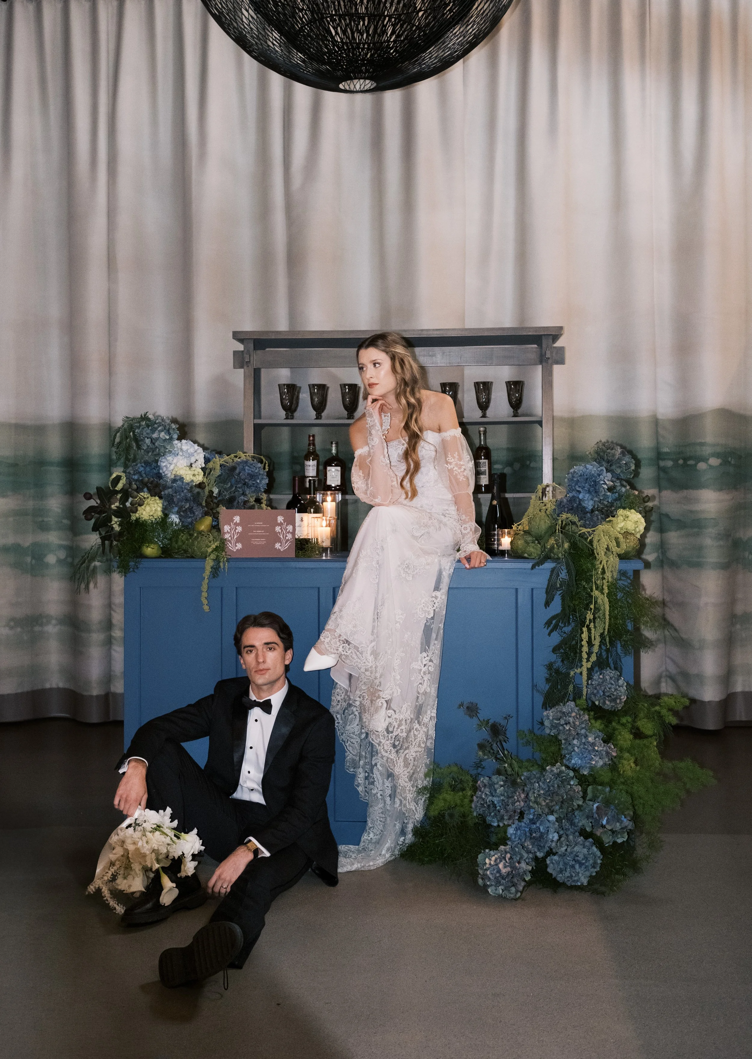 A bride and groom in wedding attire at indoor reception with floral arrangements and a blue bar behind them.
