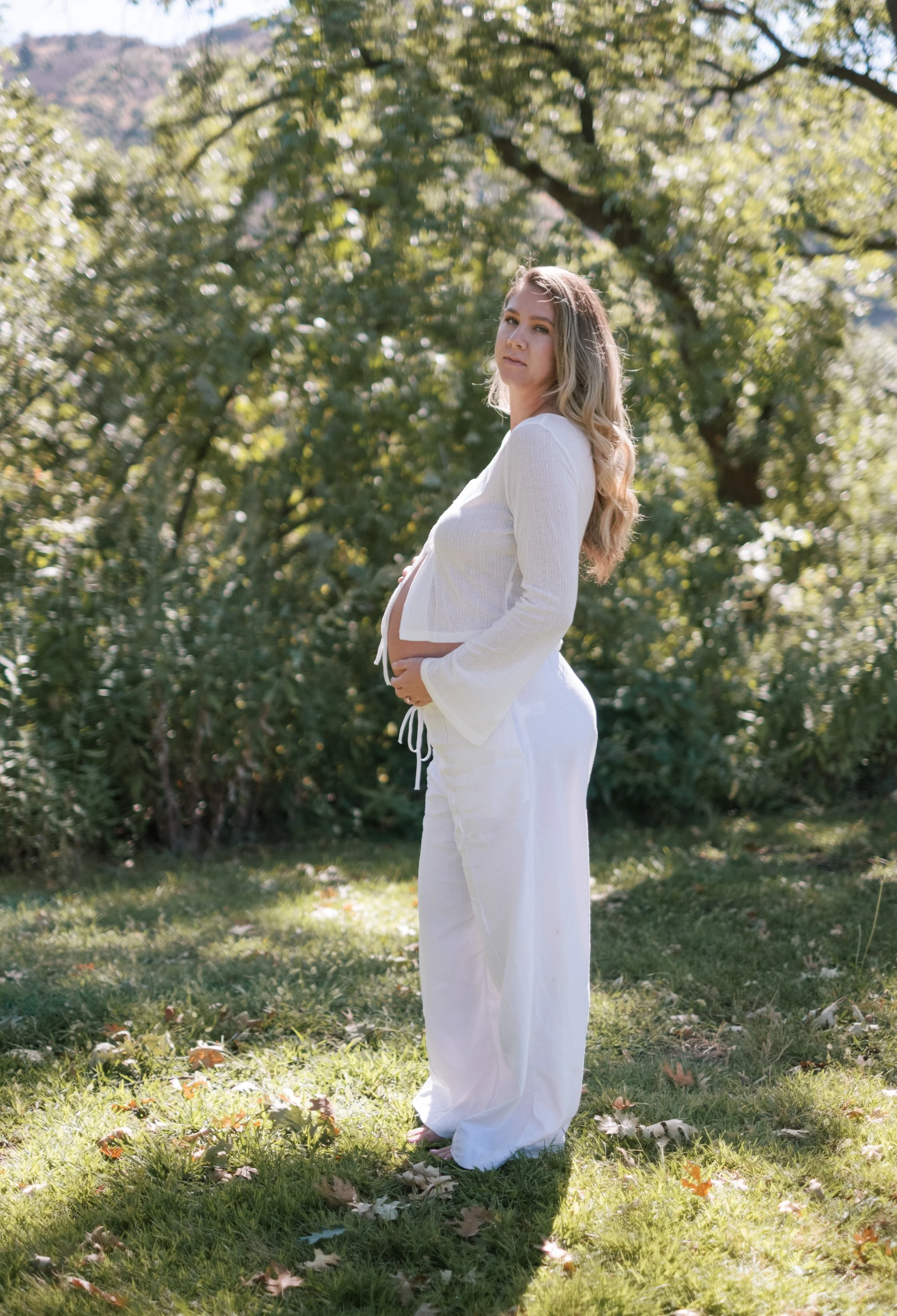 A pregnant woman in a white sweater and white pants standing outdoors on grass with fallen leaves, holding her belly, with trees and hills in the background, during daytime.