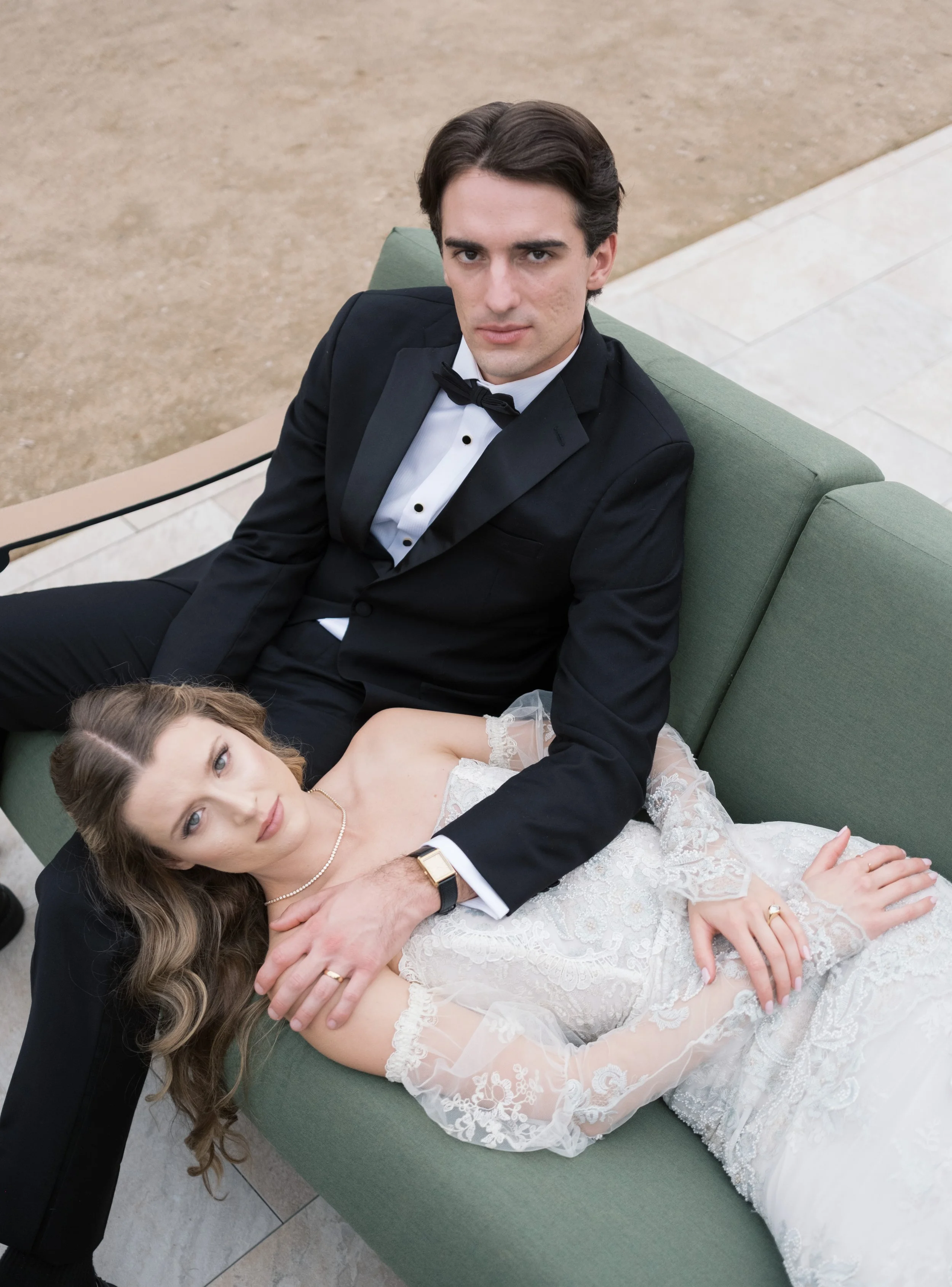 A man in a black tuxedo with a bow tie and a woman in a white lace wedding dress lying on a green sofa outdoors.