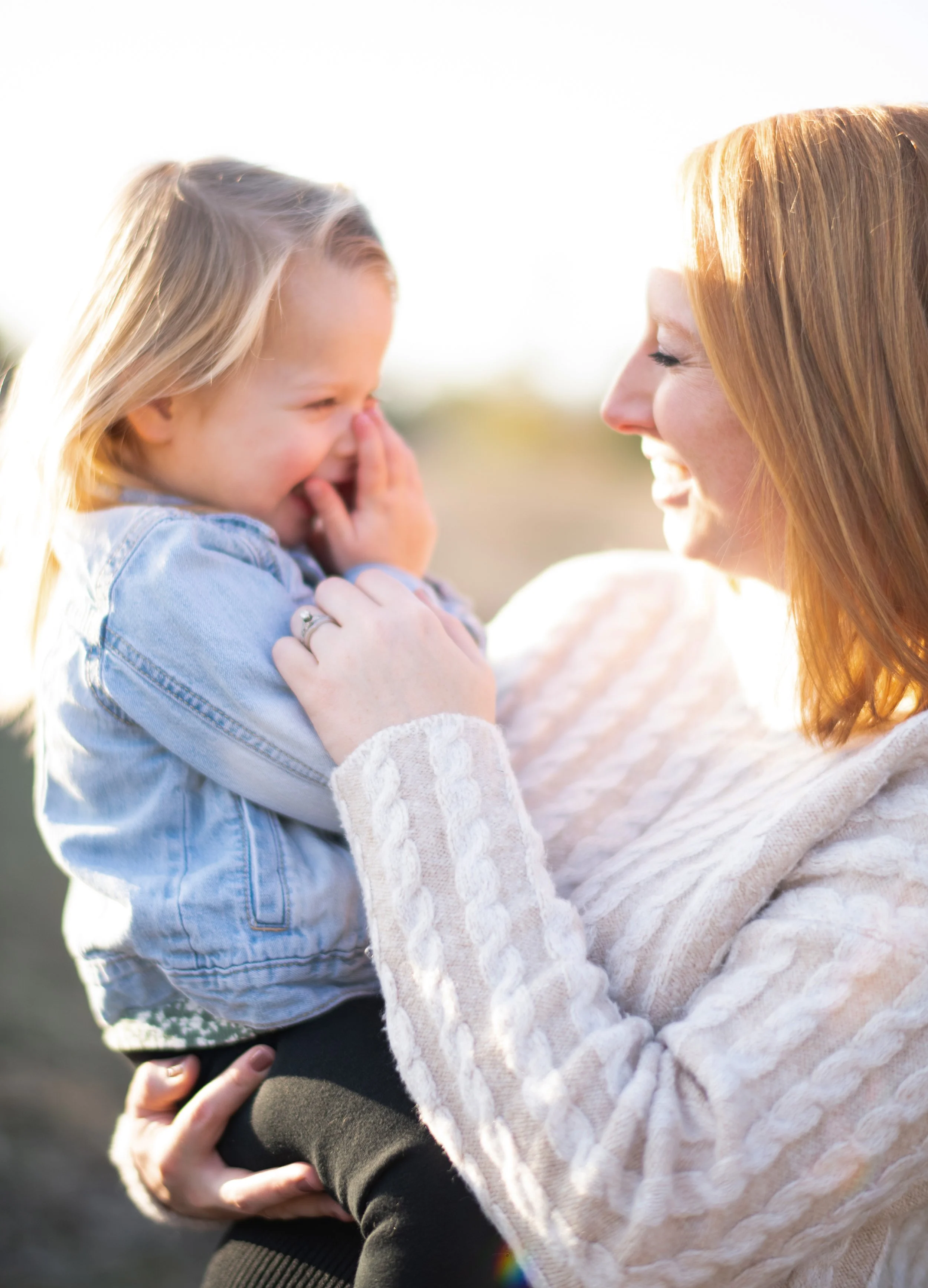 A woman and a girl are smiling and laughing while embracing outdoors. The woman has reddish hair and is wearing a white, cable-knit sweater. The girl has blonde hair and is wearing a denim jacket.