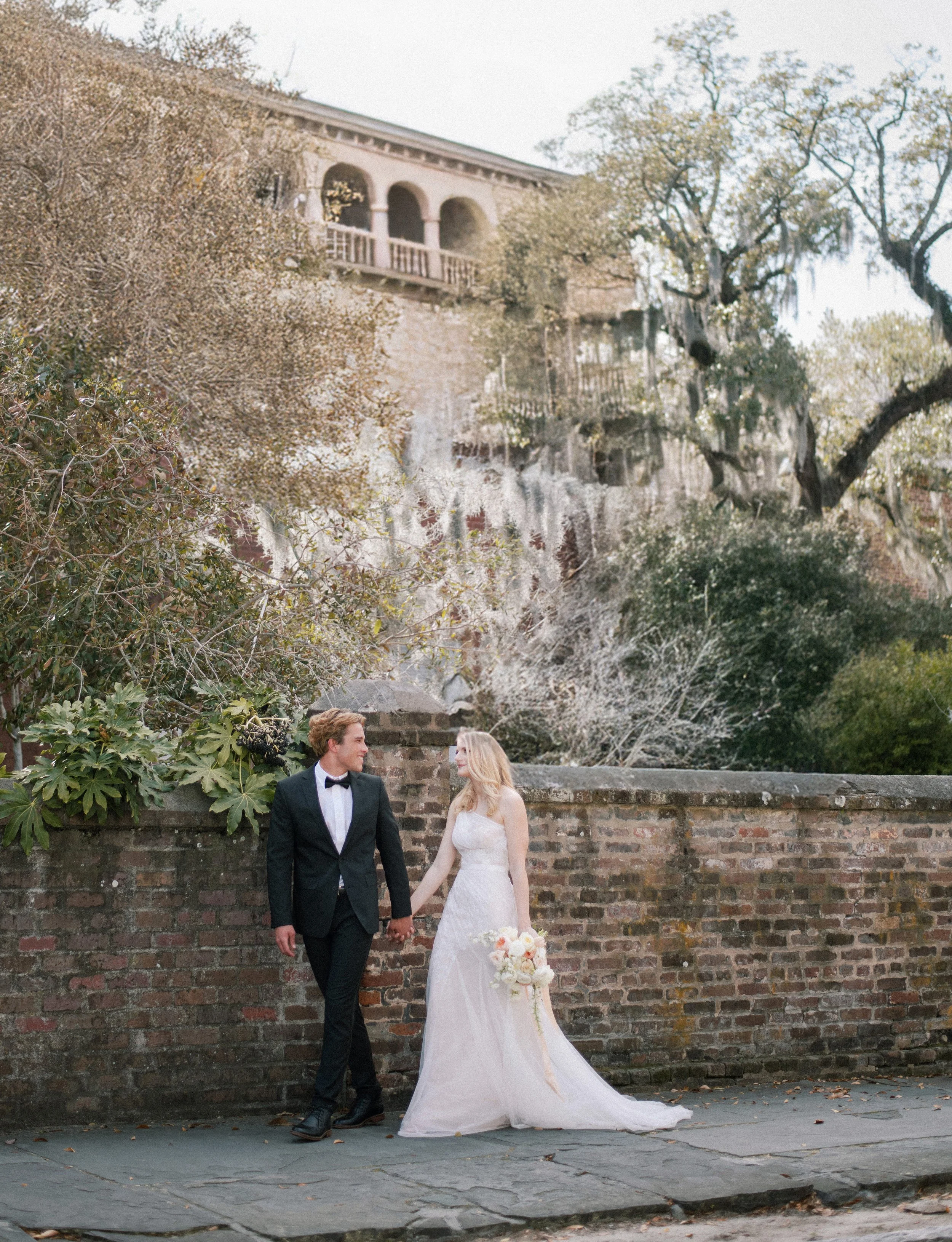 A bride and groom holding hands, standing outdoors in front of a brick wall, with trees and a building in the background. The groom is wearing a black tuxedo, and the bride is in a white wedding dress holding a bouquet of flowers.