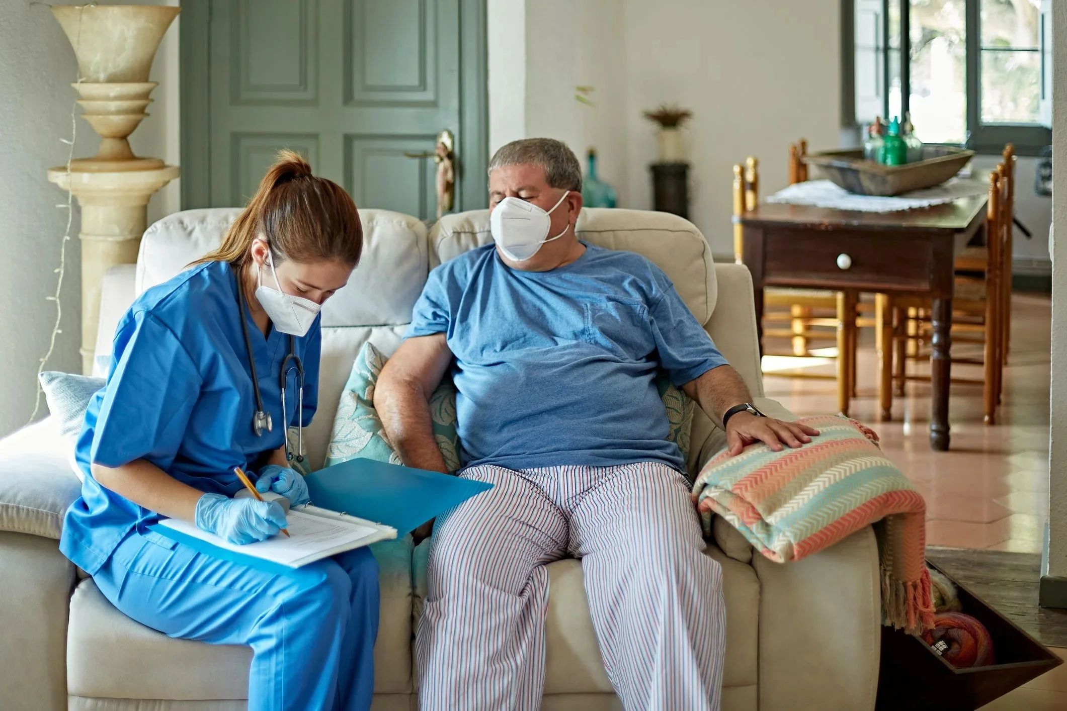 A healthcare worker and an elderly man wearing masks sitting on a couch, with the healthcare worker writing on a clipboard.