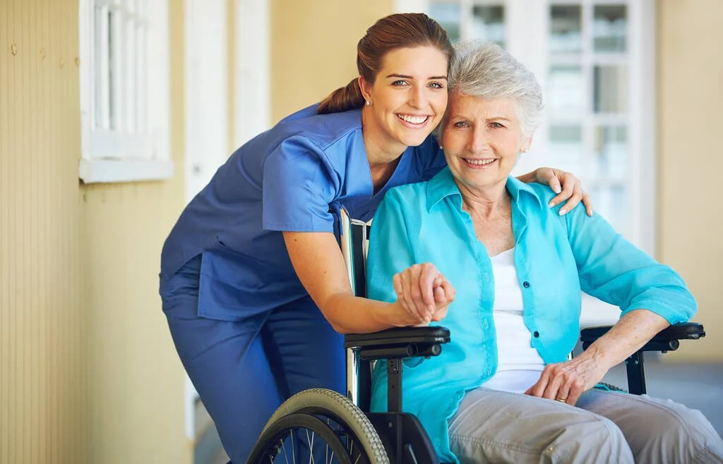A young female nurse in blue scrubs smiling and holding hands with an elderly woman in a wheelchair, in a bright indoor setting.